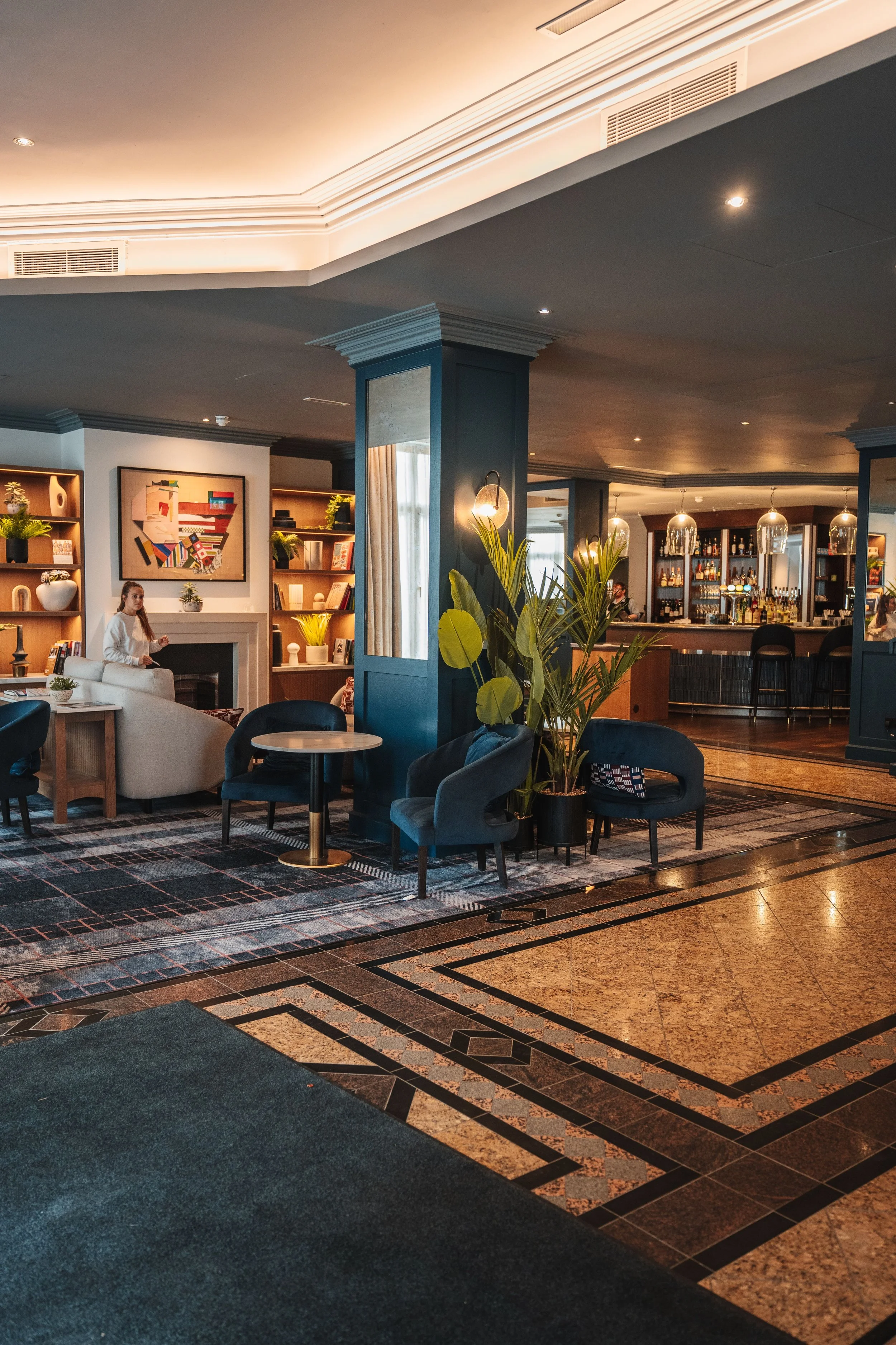 Interior of a hotel lobby with seating area, bookshelves, artwork, and a bar in the background. The lighting is warm, and there are plants and modern decor.