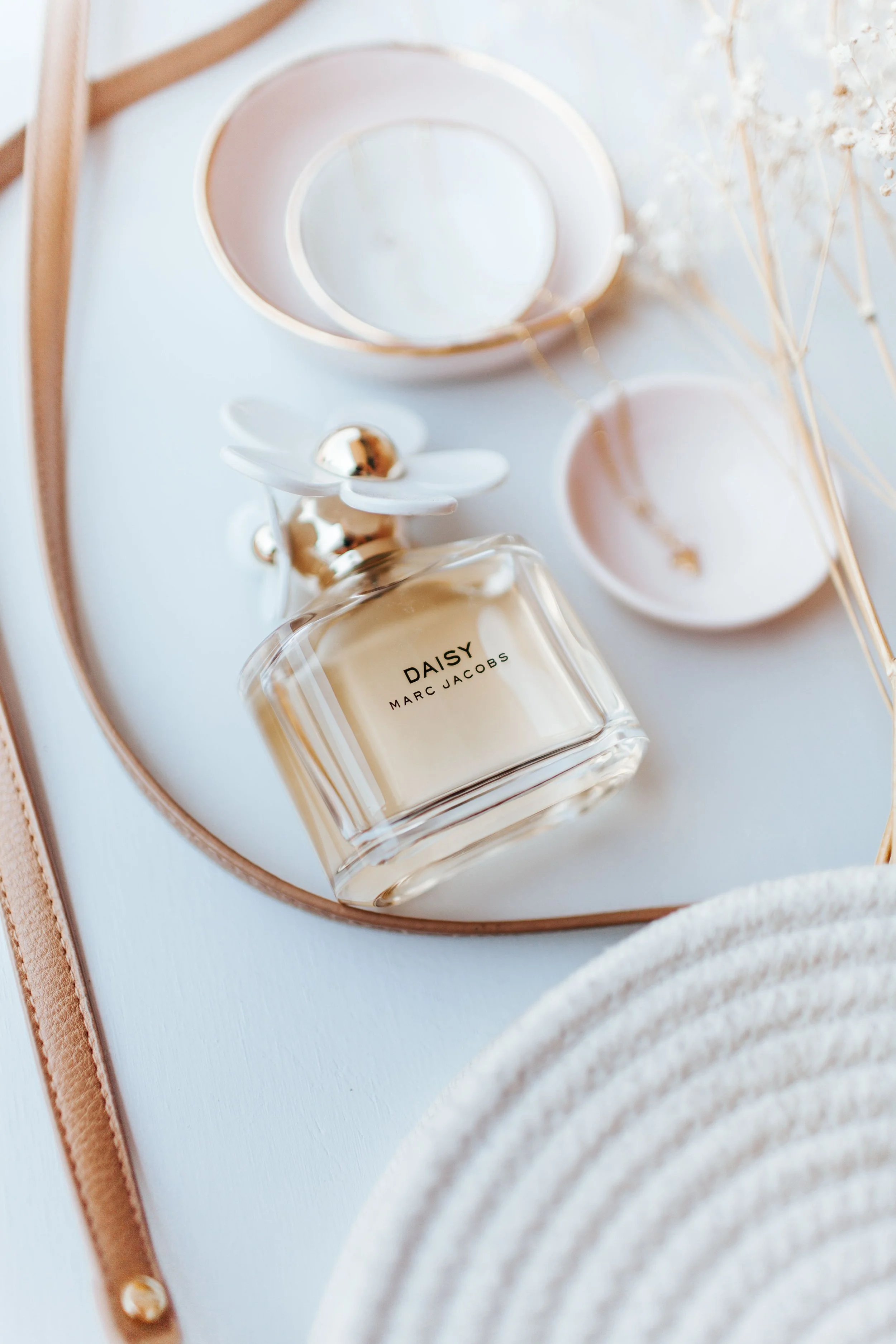 A wooden tray with a Marc Jacobs Daisy perfume bottle, two circular bowls, a white flower-shaped diffuser, and dried white flowers against a soft background.