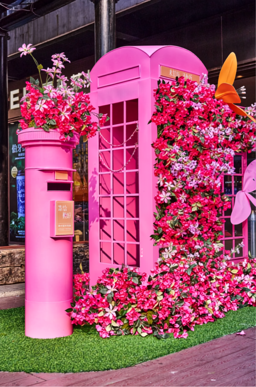 Pink telephone booth decorated with pink and white flowers, with a small pink mailbox attached, surrounded by artificial green grass.