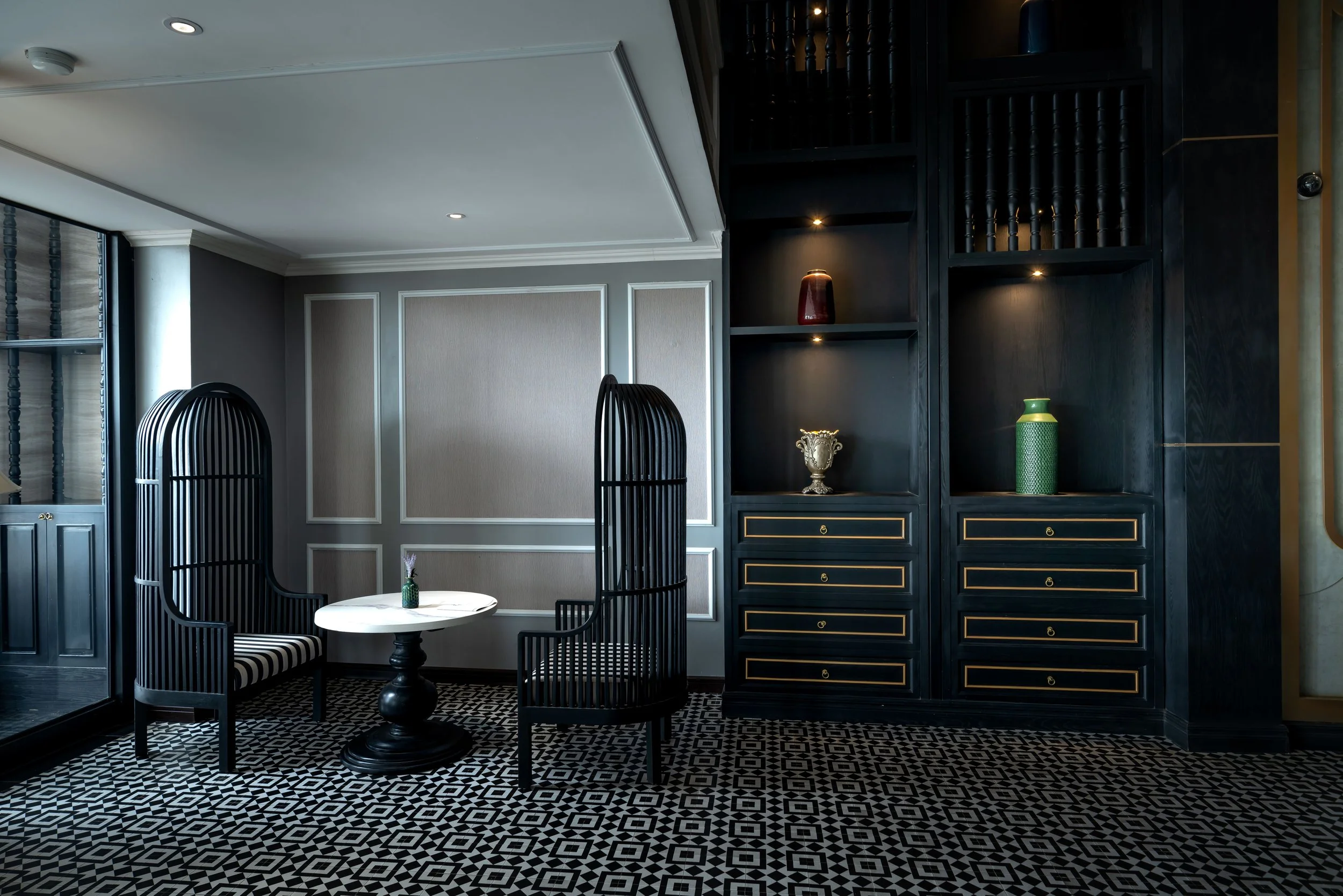 Interior of a modern room with black and white patterned tile floor, dark blue cabinetry with gold accents, black high-backed chairs with striped upholstery, and a small round table with a green vase. Light gray walls and white ceiling with crown molding.