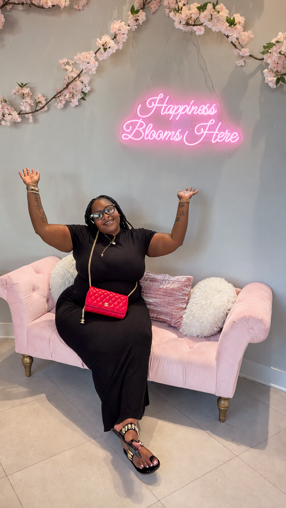 A woman sitting on a pink velvet sofa with floral pillows, smiling and raising her hands, in front of a wall with pink cherry blossom branches and a neon sign that reads 'Happiness Blooms Here'.