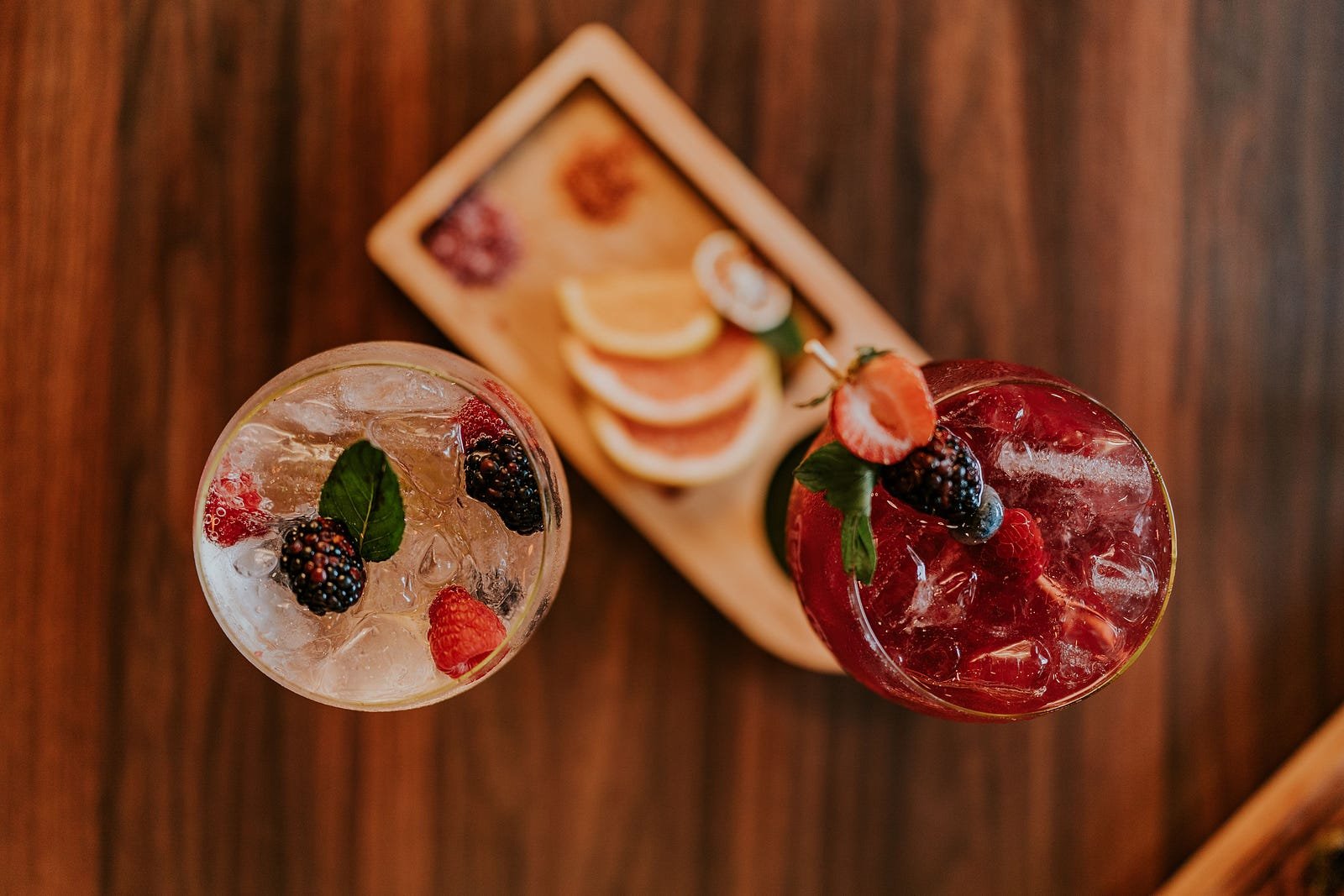 Two colorful cocktails, including a clear berry cocktail with mint and a red fruit cocktail, served with orange slices in the background.