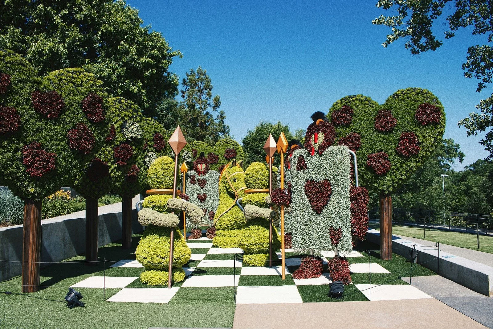 Whimsical Alice in Wonderland-inspired topiary sculptures at the Atlanta Botanical Garden, including card soldiers and heart-shaped trees along a checkered walkway.