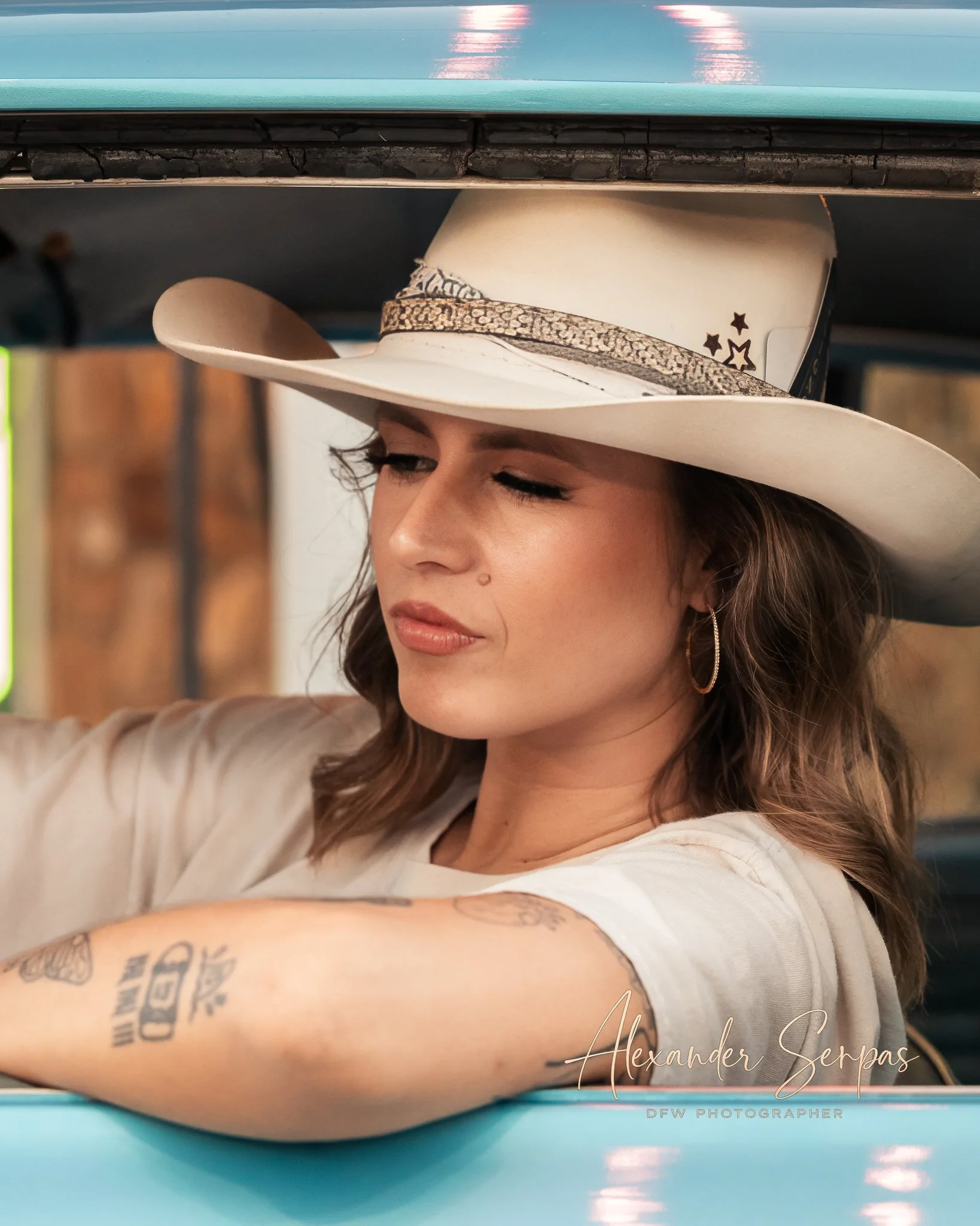 A close-up of a woman wearing a cowboy hat with star decorations. She has closed eyes and a subtle expression. The background is blurred, with some reflection on the top part of the hat. The woman's arm, with tattoos, is visible in the foreground.