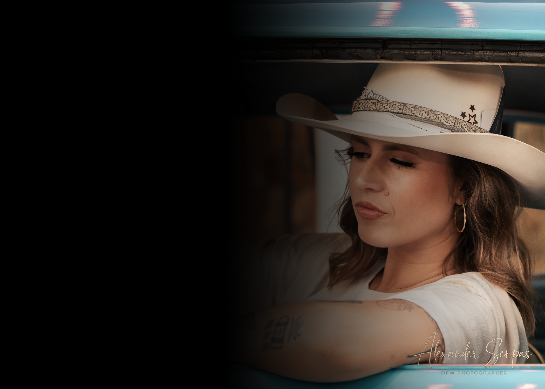 A woman with long brown hair and tattoos on her arm is wearing a white cowboy hat with star and pattern details, and is leaning against a blue surface with her eyes closed.