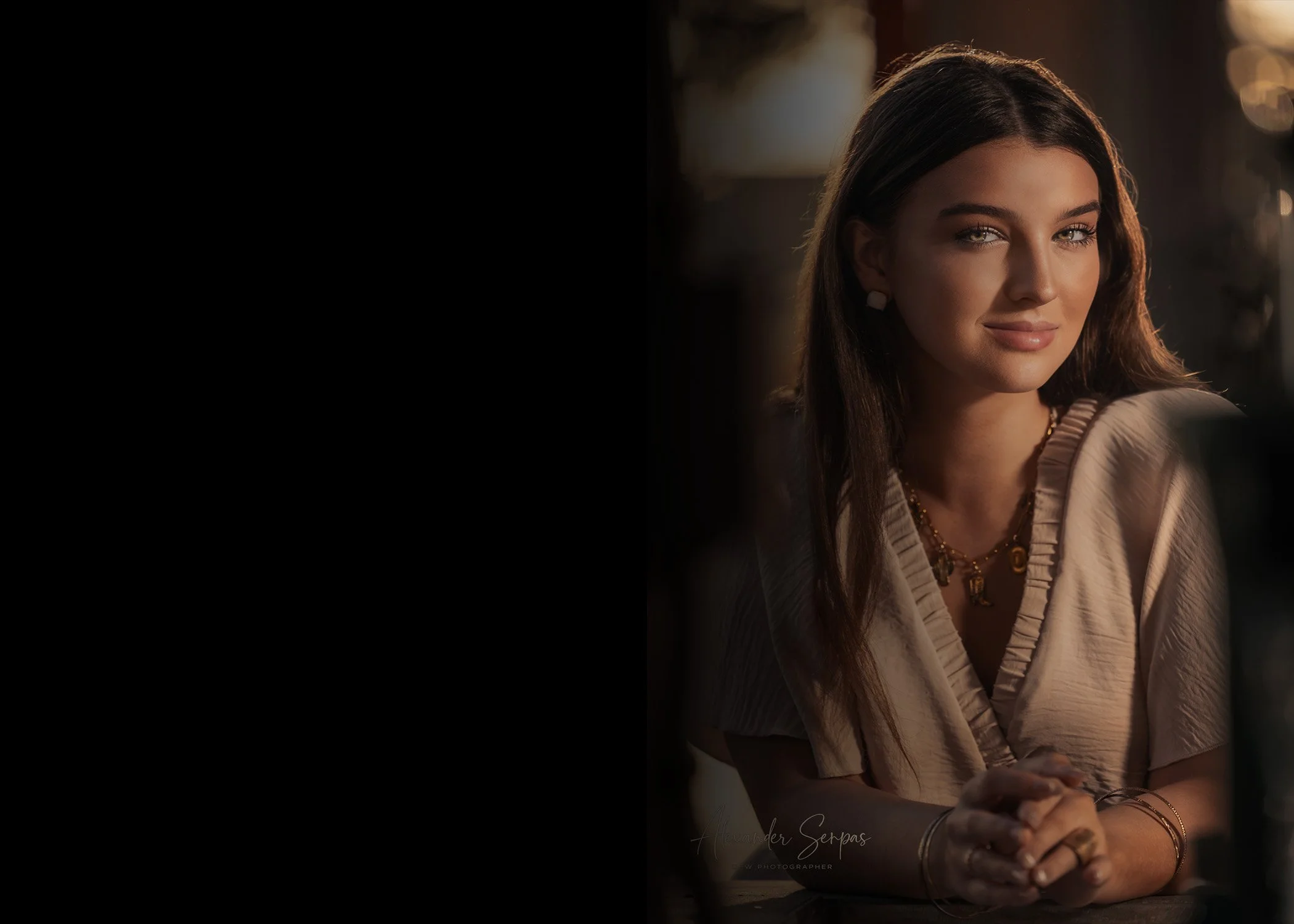 A young woman with long brown hair and green eyes, wearing a beige top, sitting indoors with warm lighting in the background.