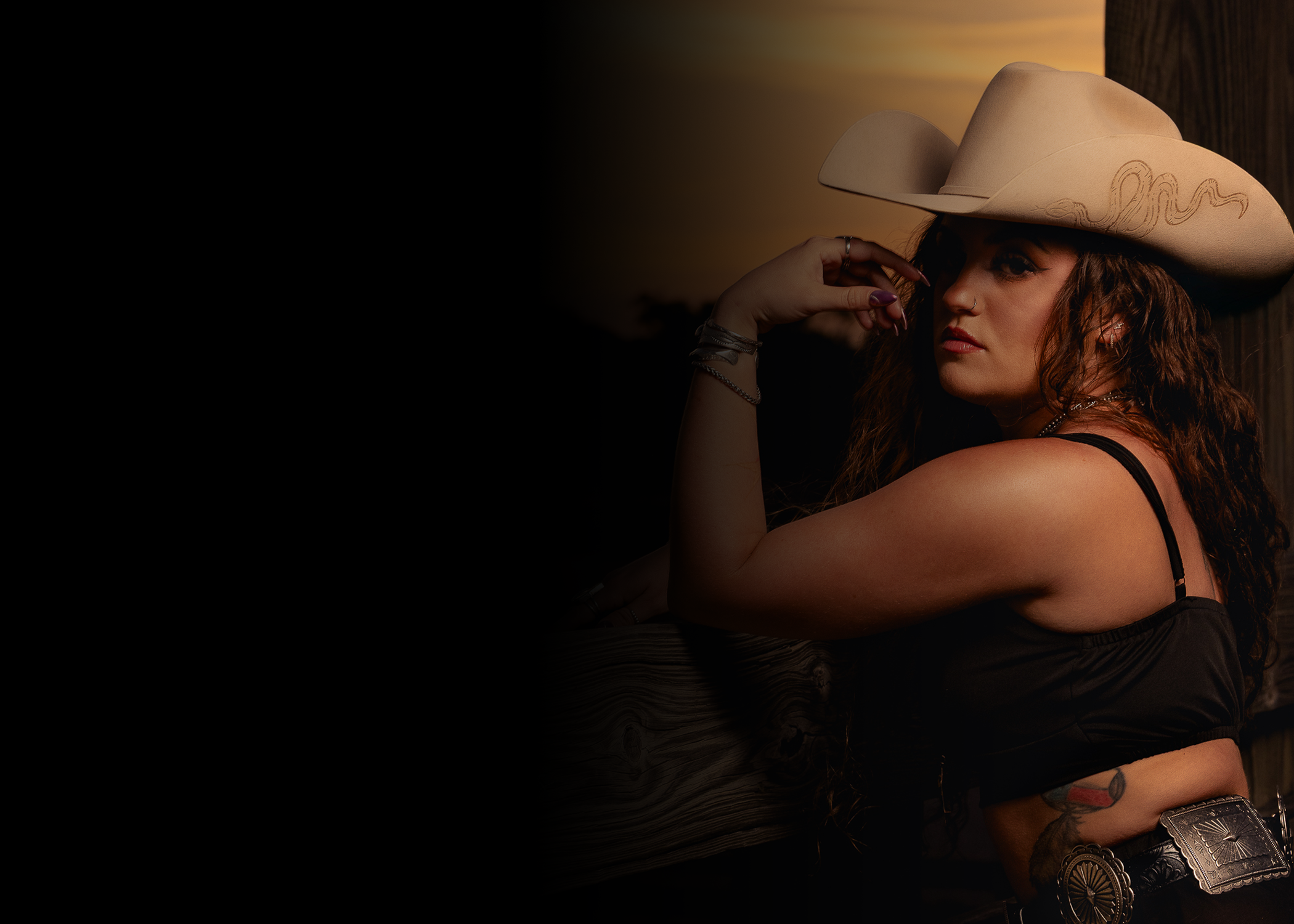 A woman with curly hair wearing a large beige cowboy hat with decorative stitching, black spaghetti straps top, and accessories, leaning her head on her hand, with sunset lighting in the background.