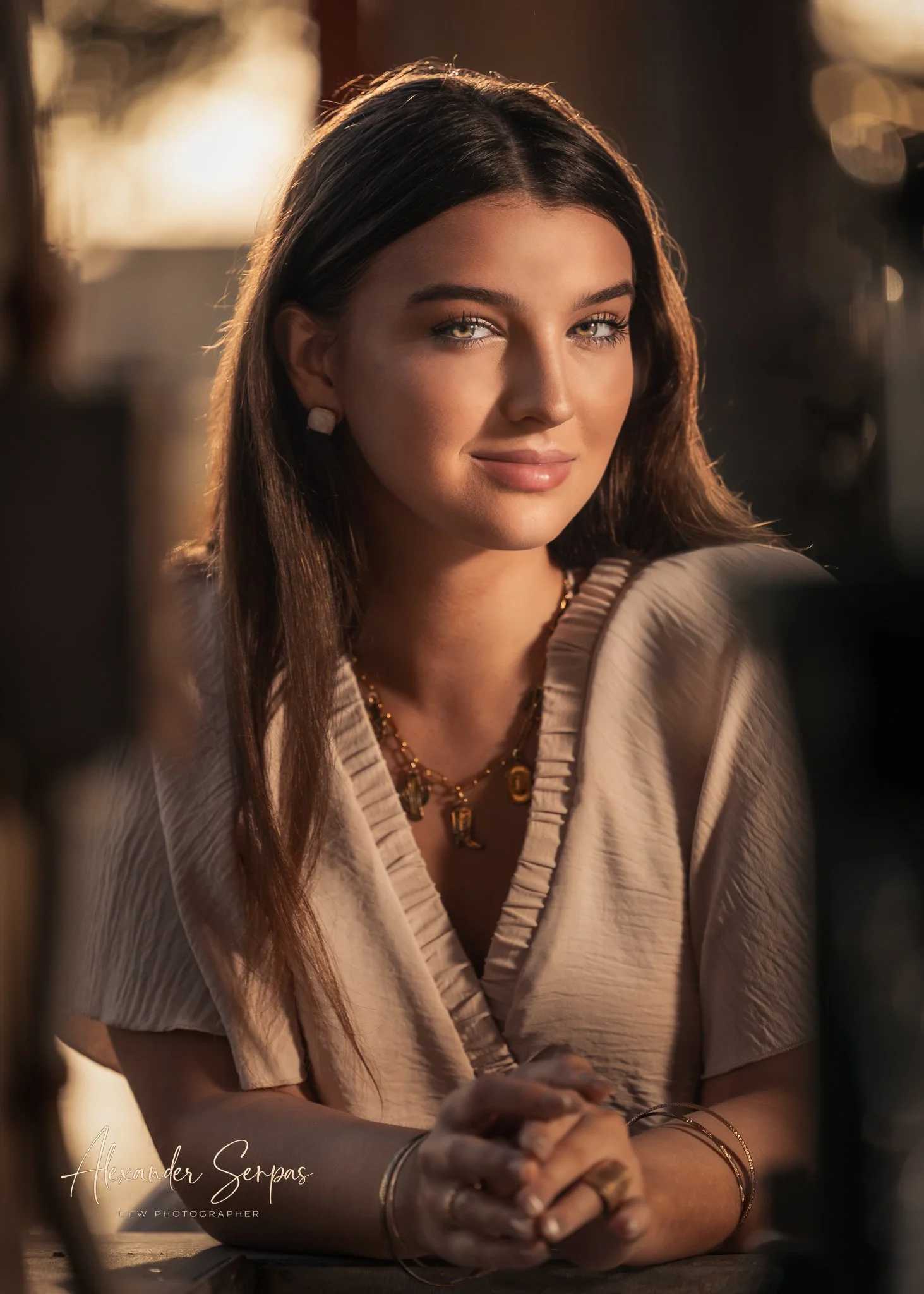 A woman with long dark hair, wearing jewelry, sits at a table with her hands folded, looking at the camera, with warm lighting and blurred background.