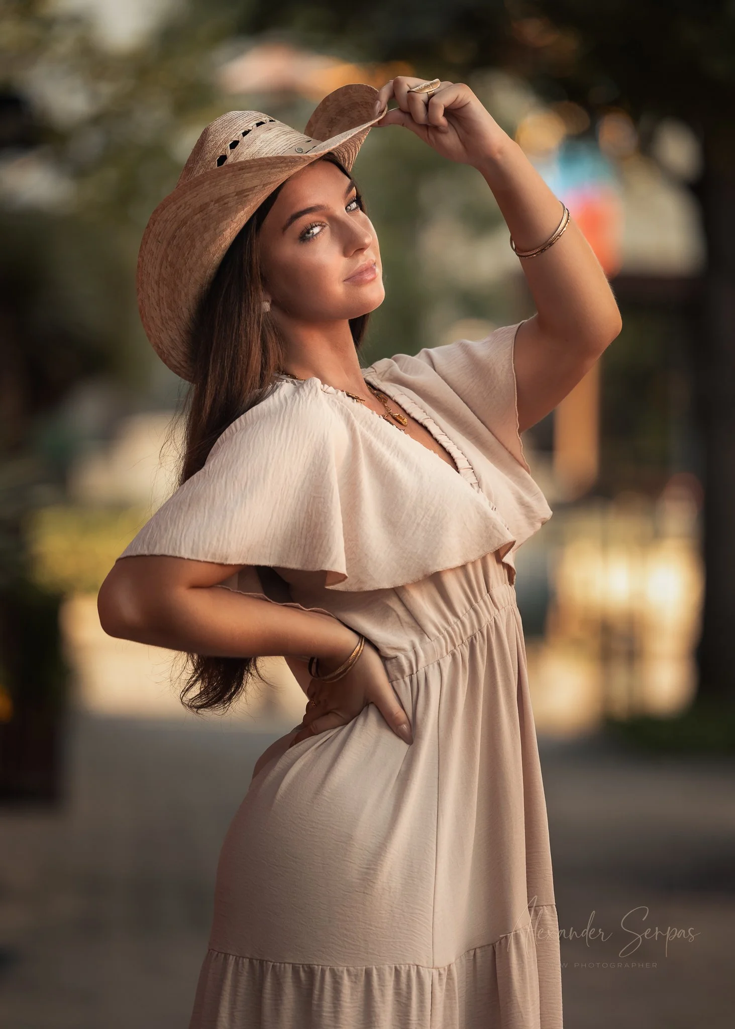 A woman wearing a large straw hat and a beige dress, standing outdoors with a blurry background of trees and water, looking confidently at the camera.