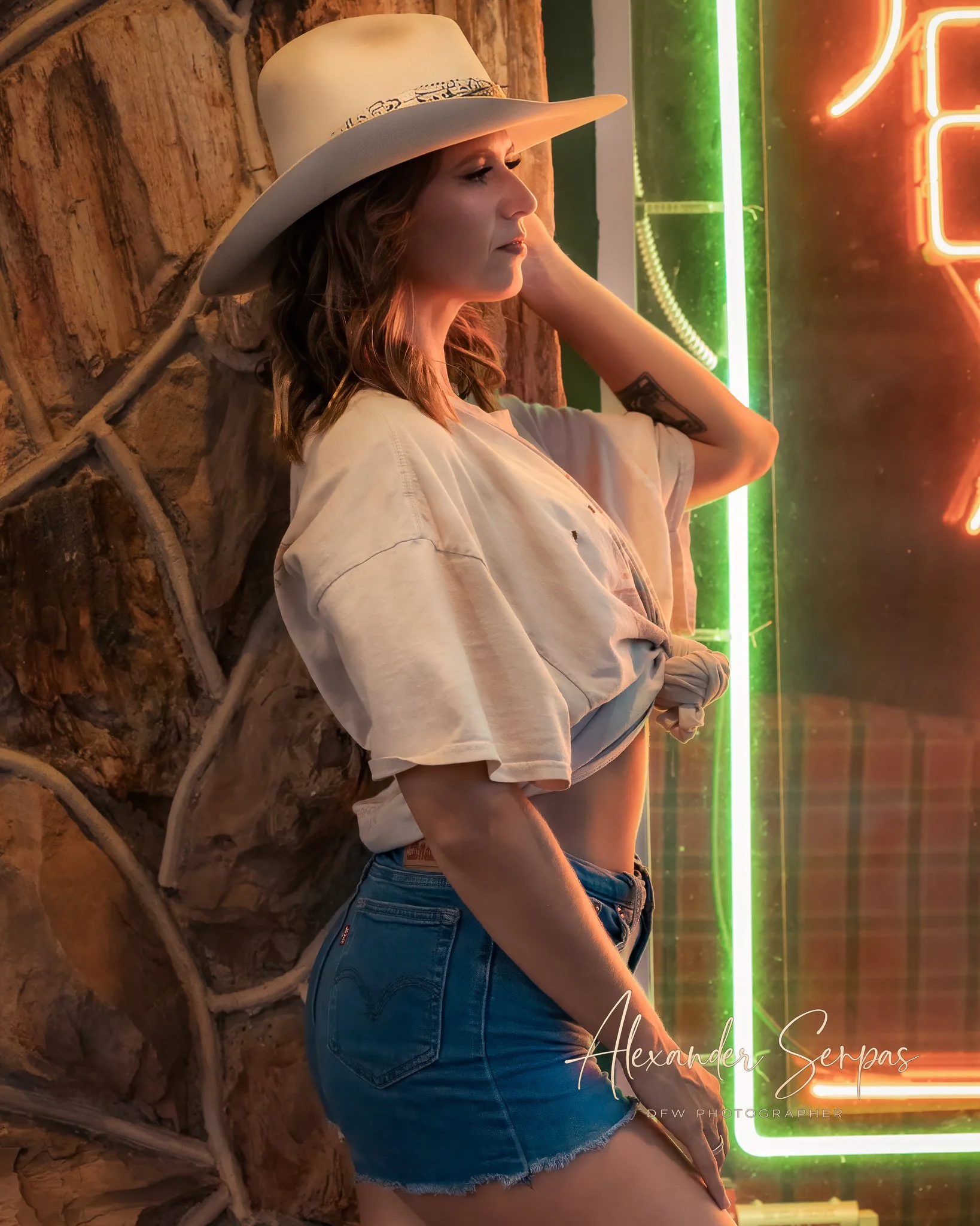 A woman with wavy hair wearing a wide-brimmed hat and a beige t-shirt, standing sideways against a rustic wooden wall with climbing plants, illuminated by colorful neon lights.