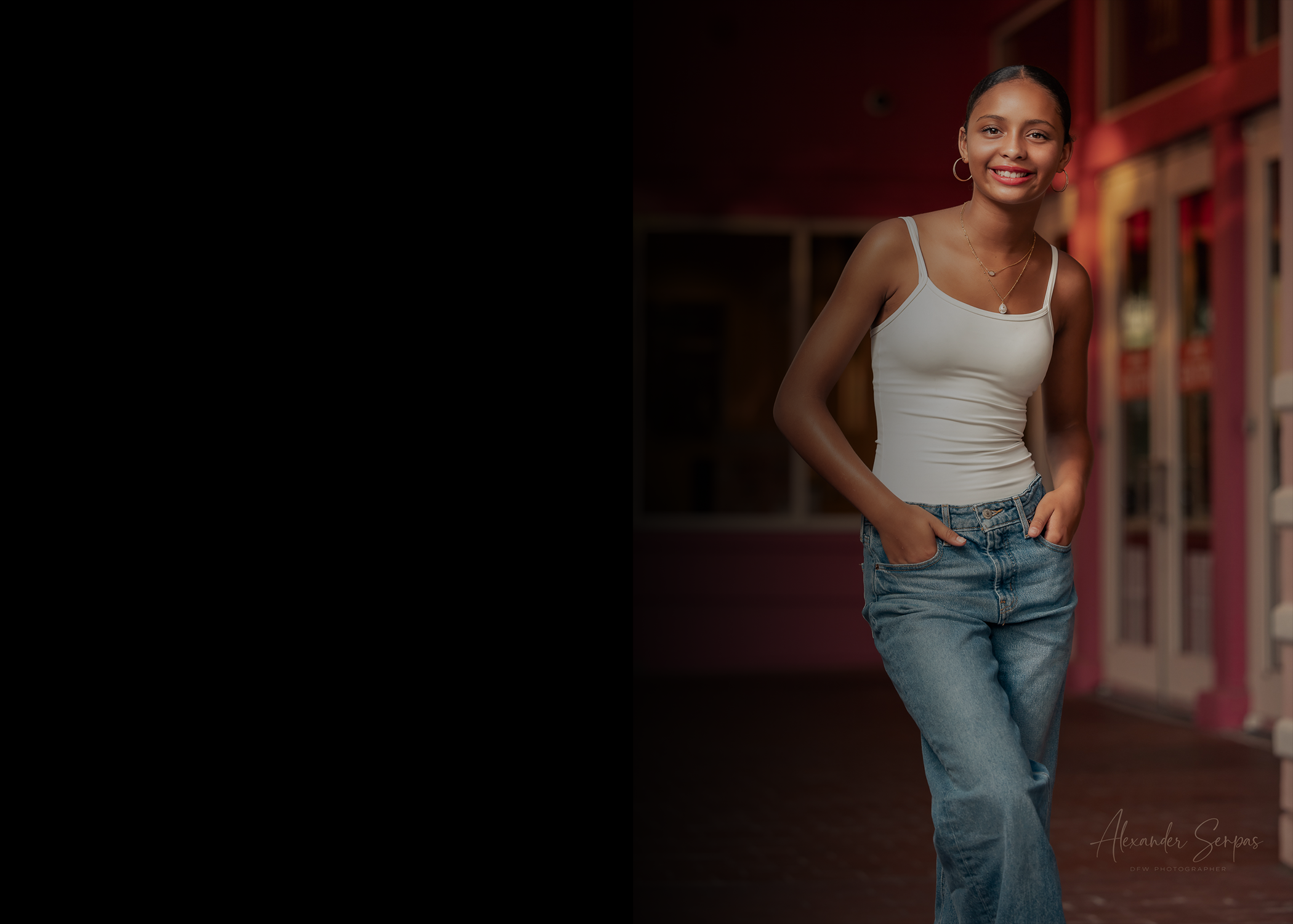 A young woman with dark hair styled in a bun, smiling and standing with her hands in the pockets of her blue jeans. She is wearing hoop earrings, layered necklaces, and a white tank top. The background features a colorful street scene with blurred storefronts and buildings.