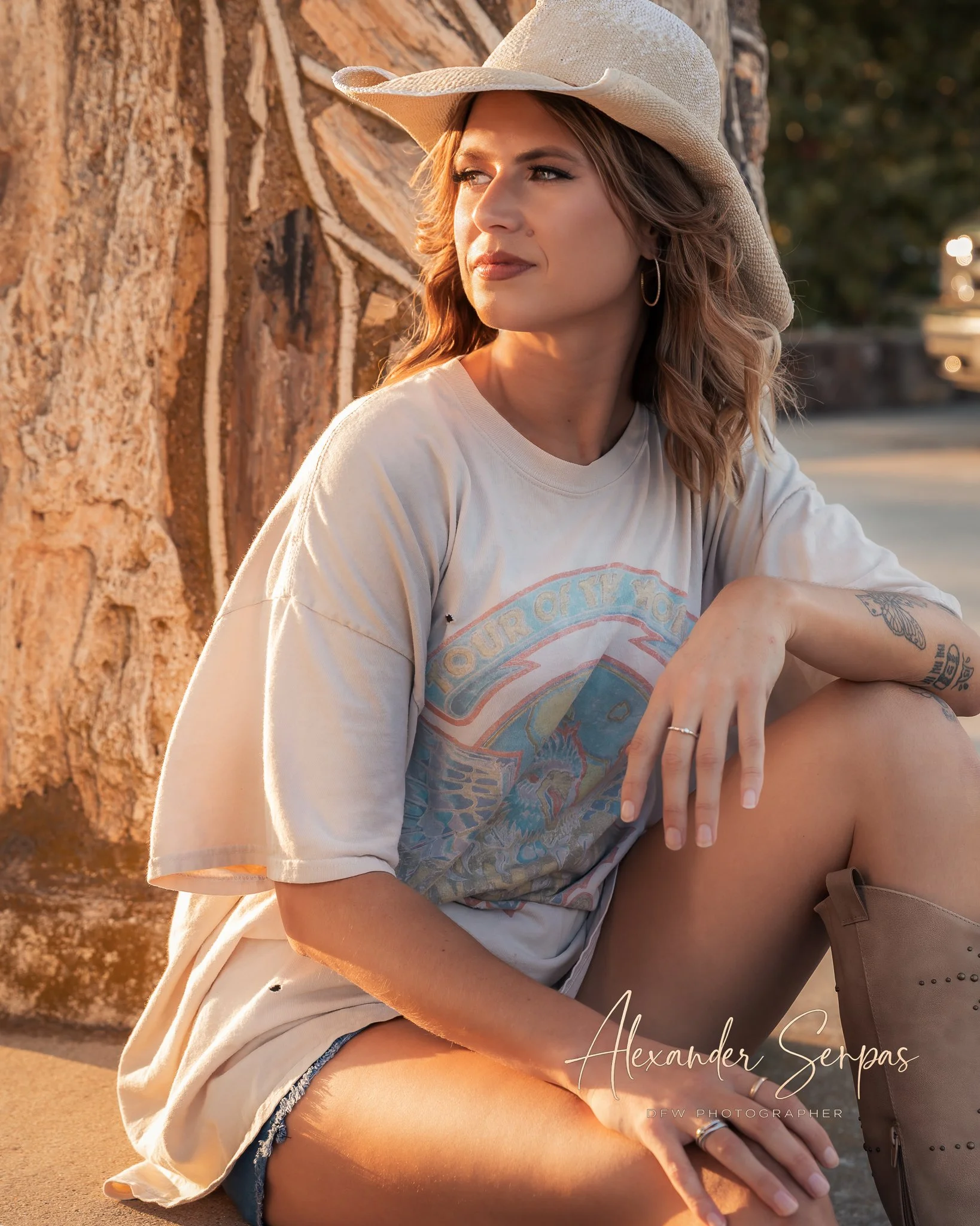 A young woman with long hair wearing a large straw hat, a vintage t-shirt, and denim shorts, sitting outdoors near a tree trunk during sunset, with a thoughtful expression, crossed arms, and tattoos on her arm.