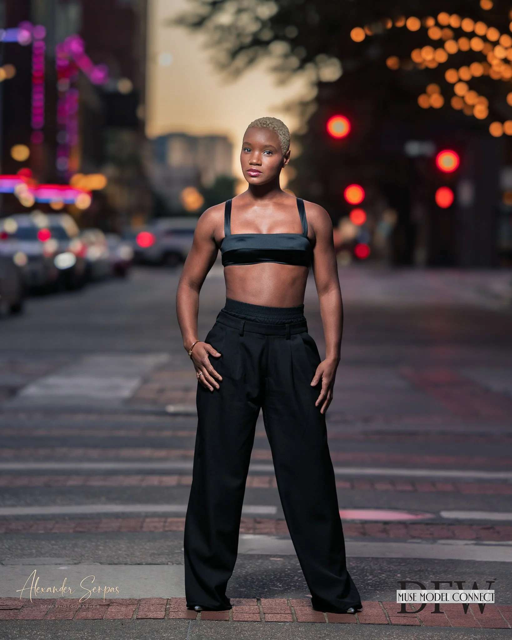 A woman with short blonde hair standing on a city street during dusk, wearing a black crop top and wide-leg black trousers, with blurred city lights and cars in the background.