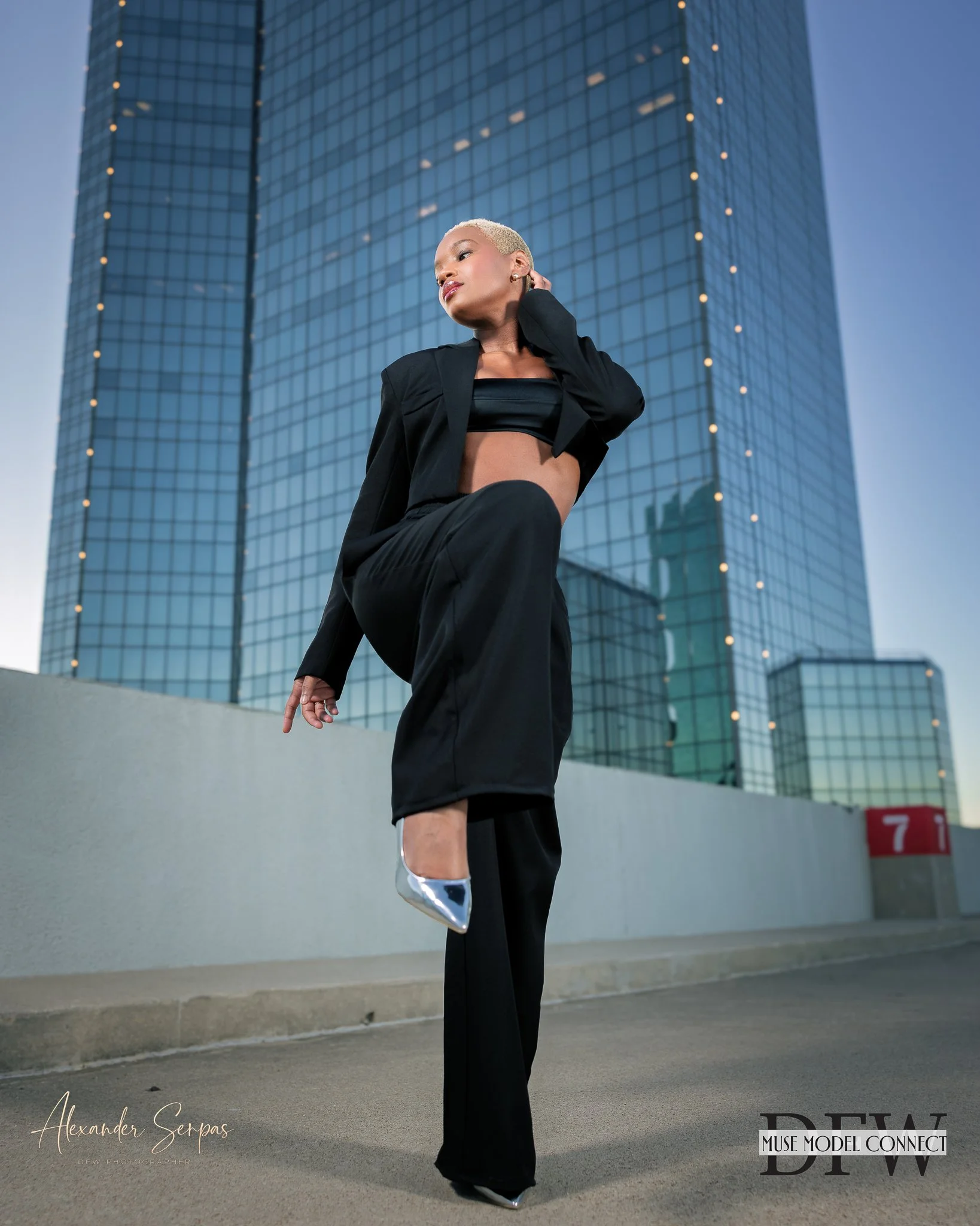 Fashion model with short blonde hair wearing a black crop top and wide-leg pants, posing outdoors in front of a tall glass office building during dusk, with text overlay for a photoshoot.