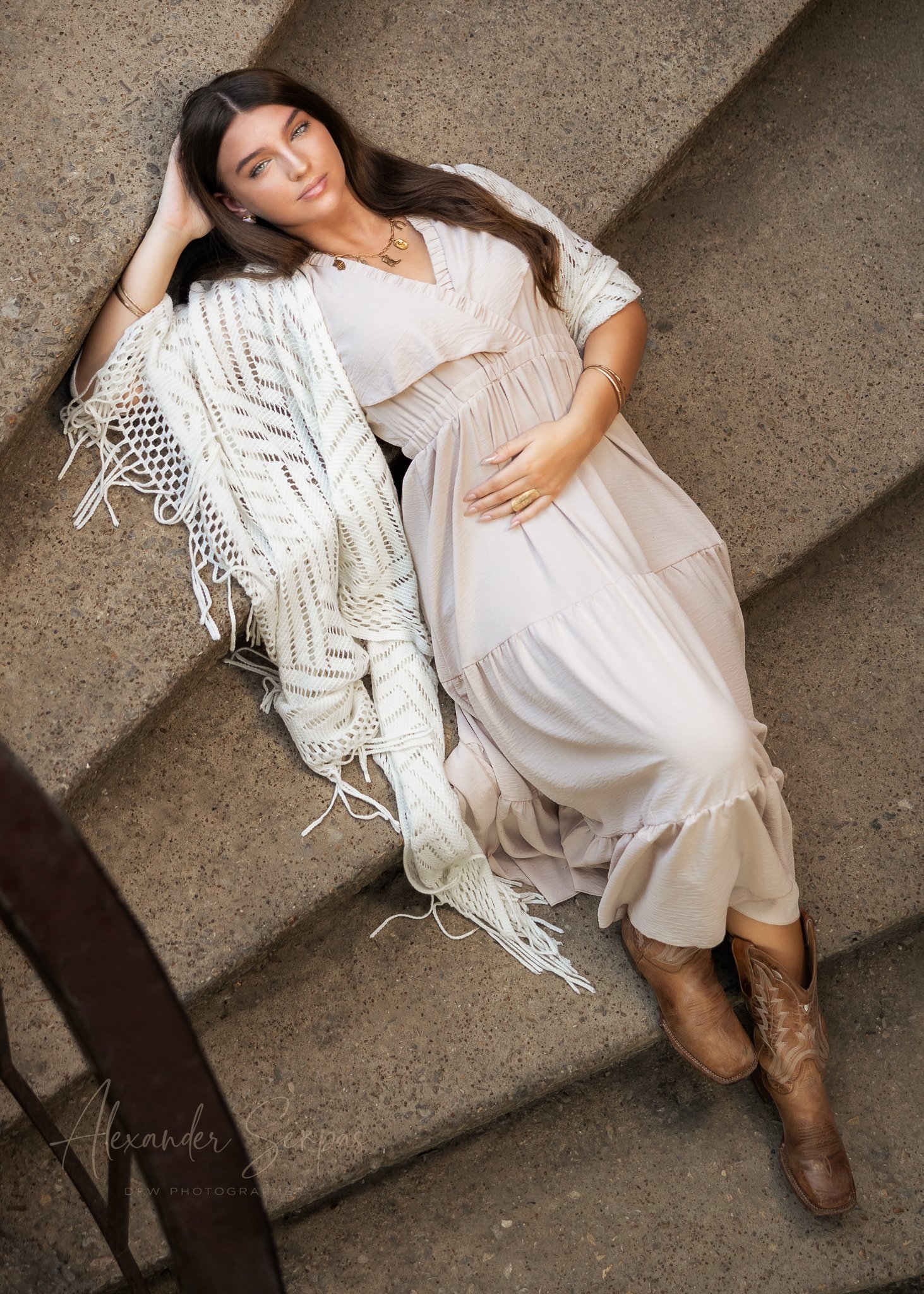 A young woman lying on a concrete step, dressed in a light-colored dress and western boots, with jewelry, resting her head on her hand and gazing at the camera.