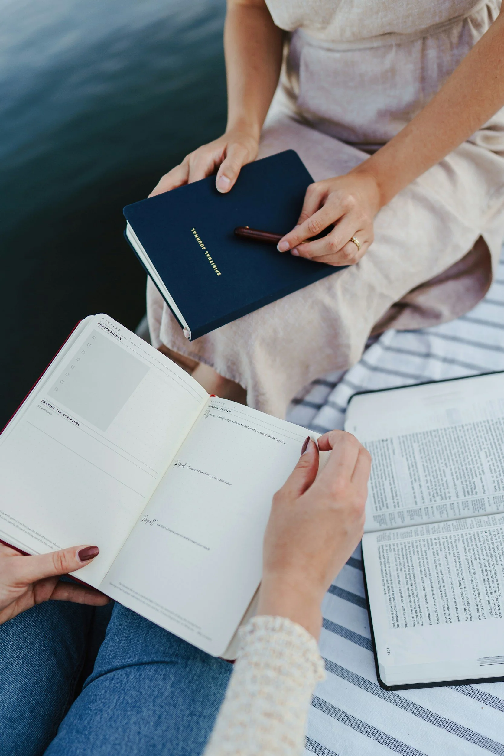 People reading a Bible and a notebook outdoors on a striped tablecloth by the water.