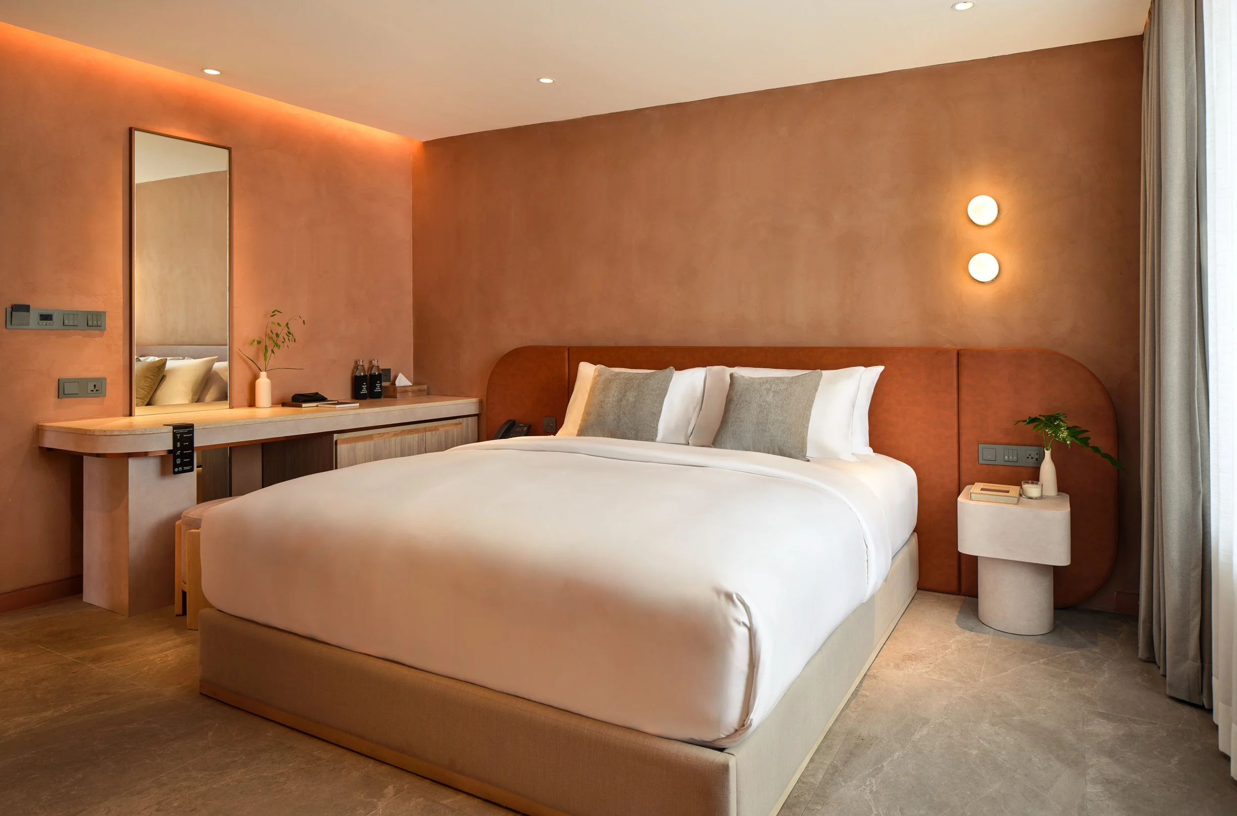 A modern hotel room with a large bed, beige headboard, white bedding, and pillows. There is a small bedside table with a plant, a telephone, and some water bottles. A mirror and some lighting fixtures are visible on the wall, and there is a curtain b