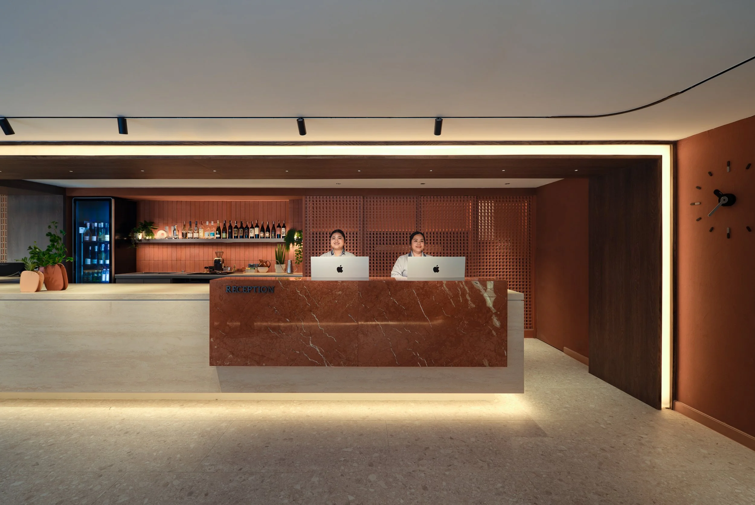 Modern hotel reception area with two female receptionists sitting at a marble counter with Apple iMacs, behind a pink brick wall bar with bottles and plants, and a large round clock on the wall.