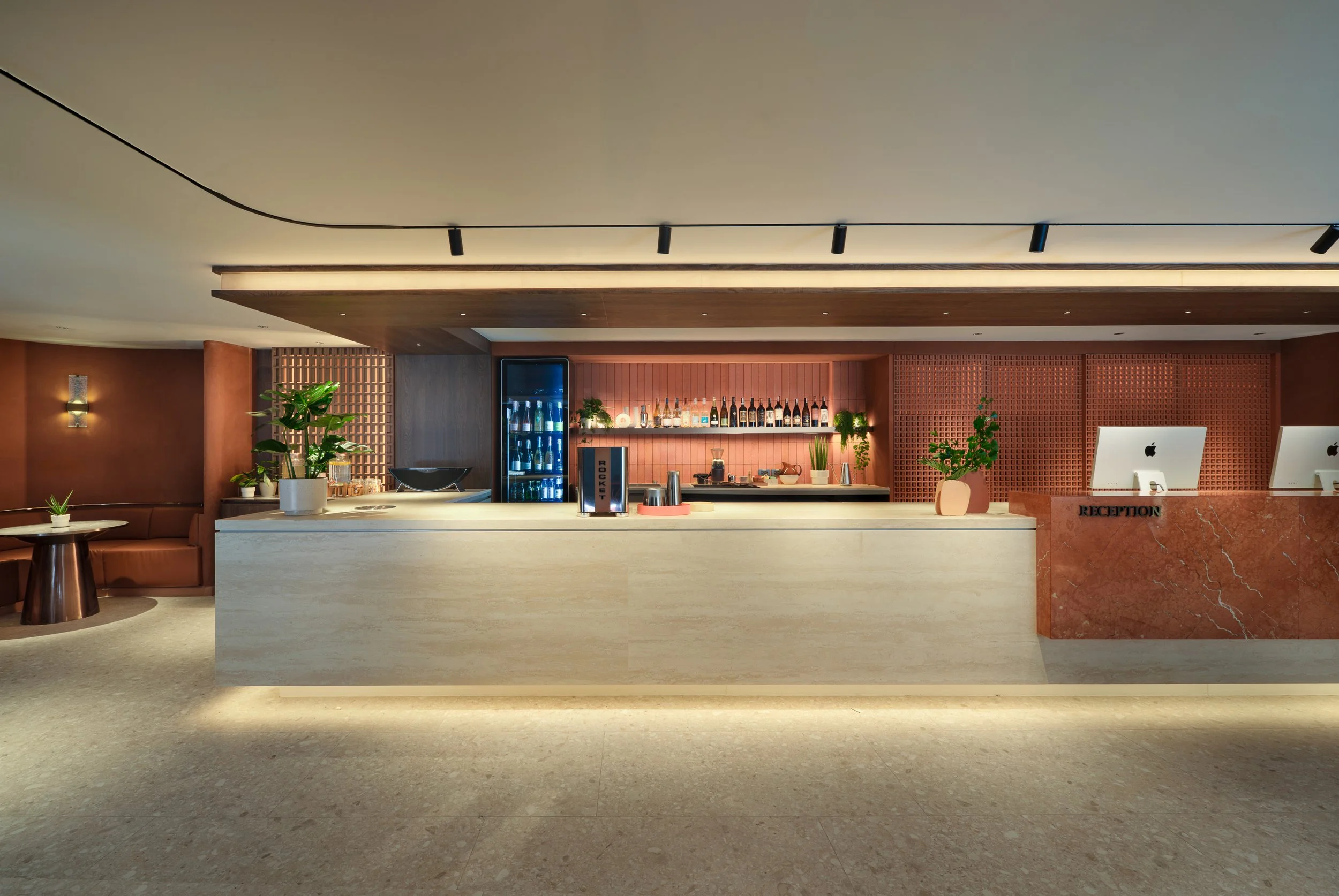 Hotel reception area with a marble front desk, potted plants, computer screens, and a display cabinet with bottles of alcohol against a wooden backdrop.