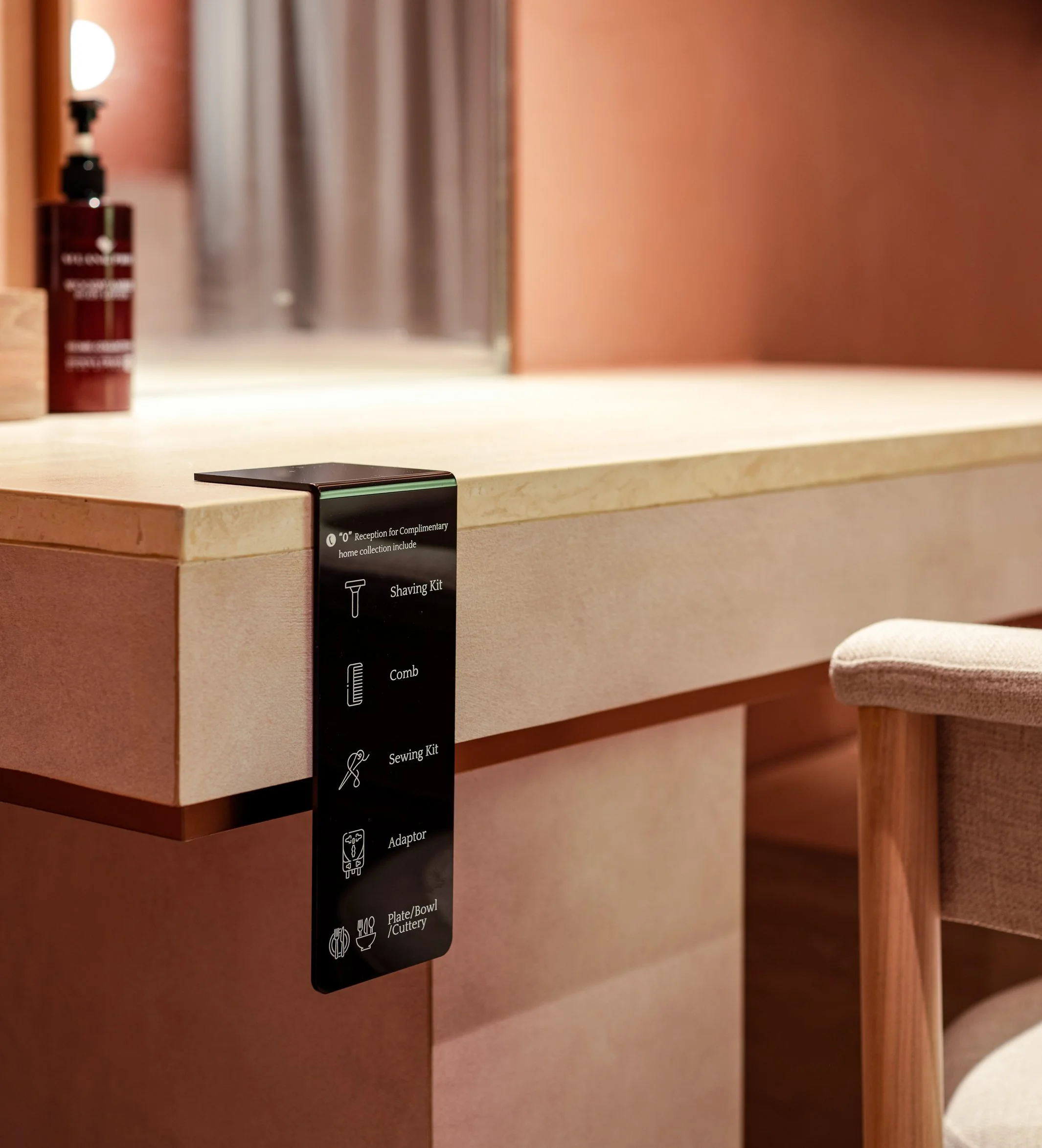 Close-up of a beige countertop with a black label indicating various hotel amenities such as a shaving kit, comb, adapter, and plate/bowl/cutlery. A partial view of a stool and a bottle of skincare product are in the background.