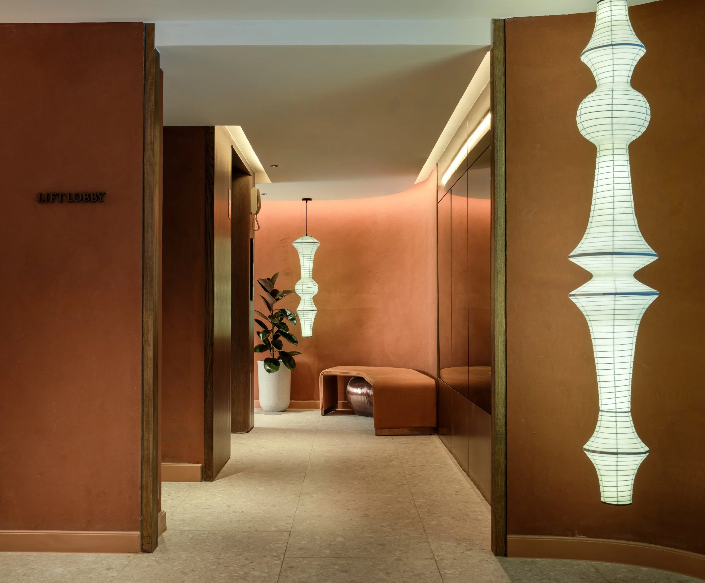 View of a hallway with wall-mounted lamps, a potted plant, and wooden partitions, illuminated by warm lights.