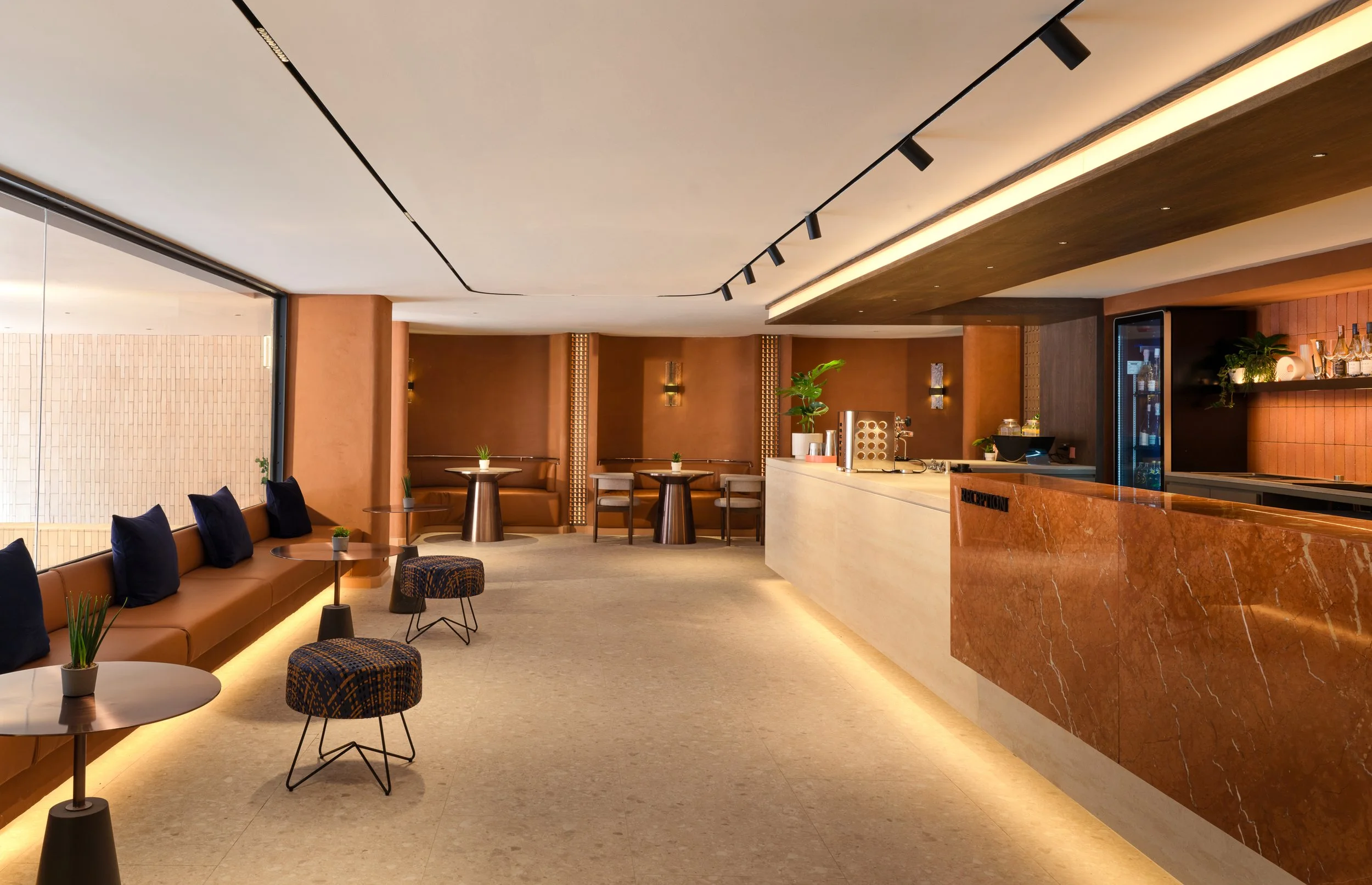 Modern hotel lobby with seating area, small tables, plants, warm lighting, and a front desk with a marble counter.