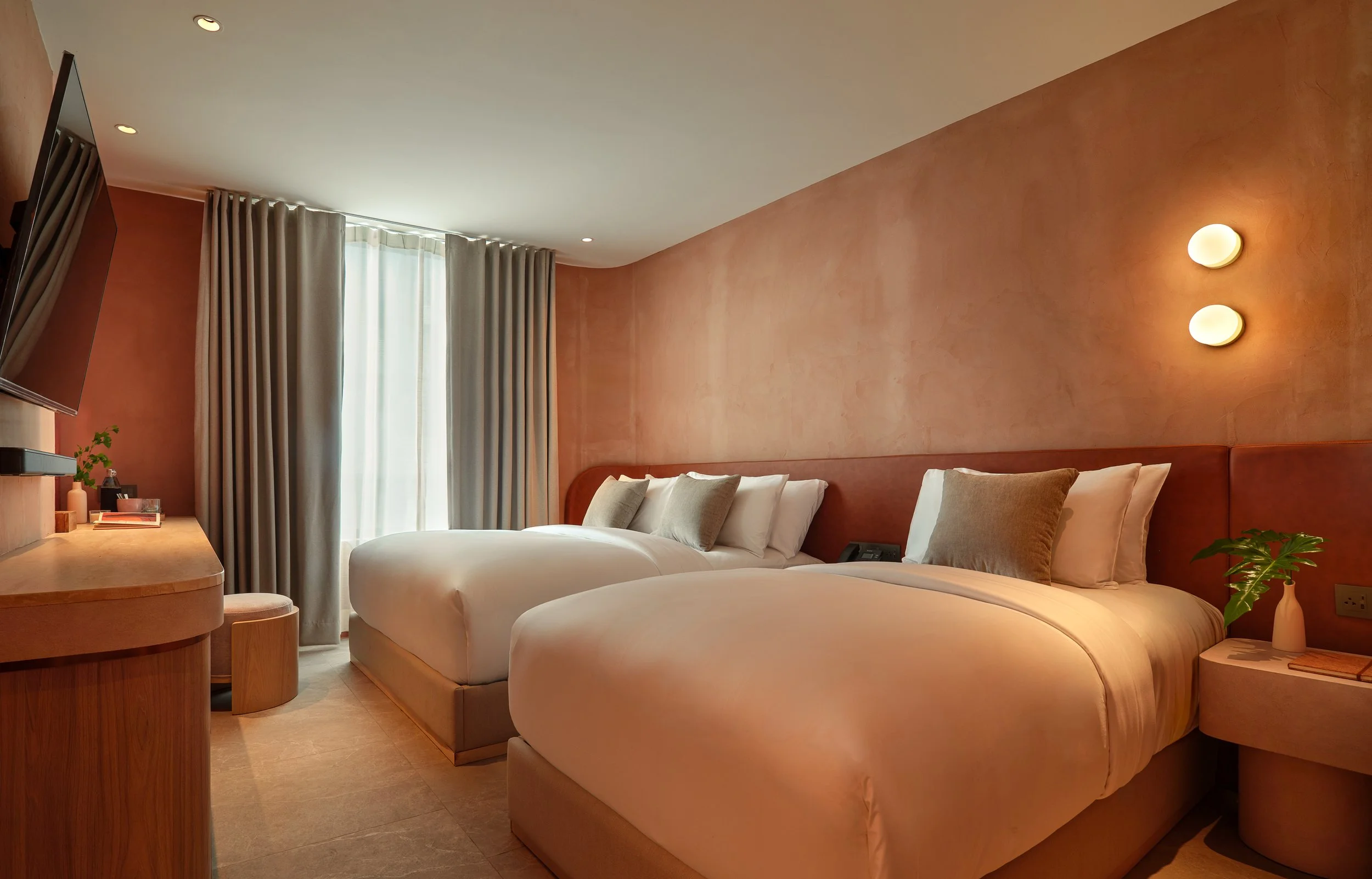 A cozy hotel room with two beds, a wooden desk, and a large window with beige curtains. The room has warm earth-tone walls and soft lighting.