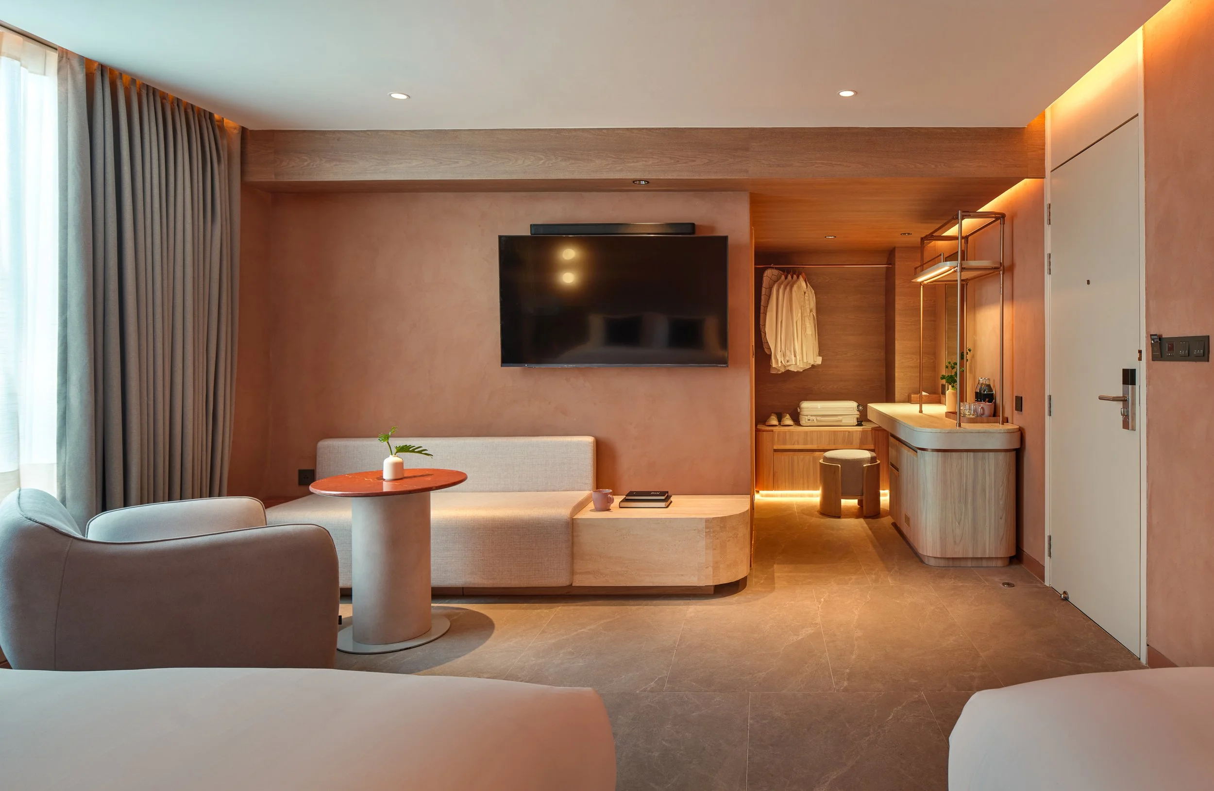 Modern hotel room with beige furniture, a wall-mounted TV, a small round table with a plant, a built-in wardrobe, and a vanity area with a mirror and shelving, illuminated with warm lighting.