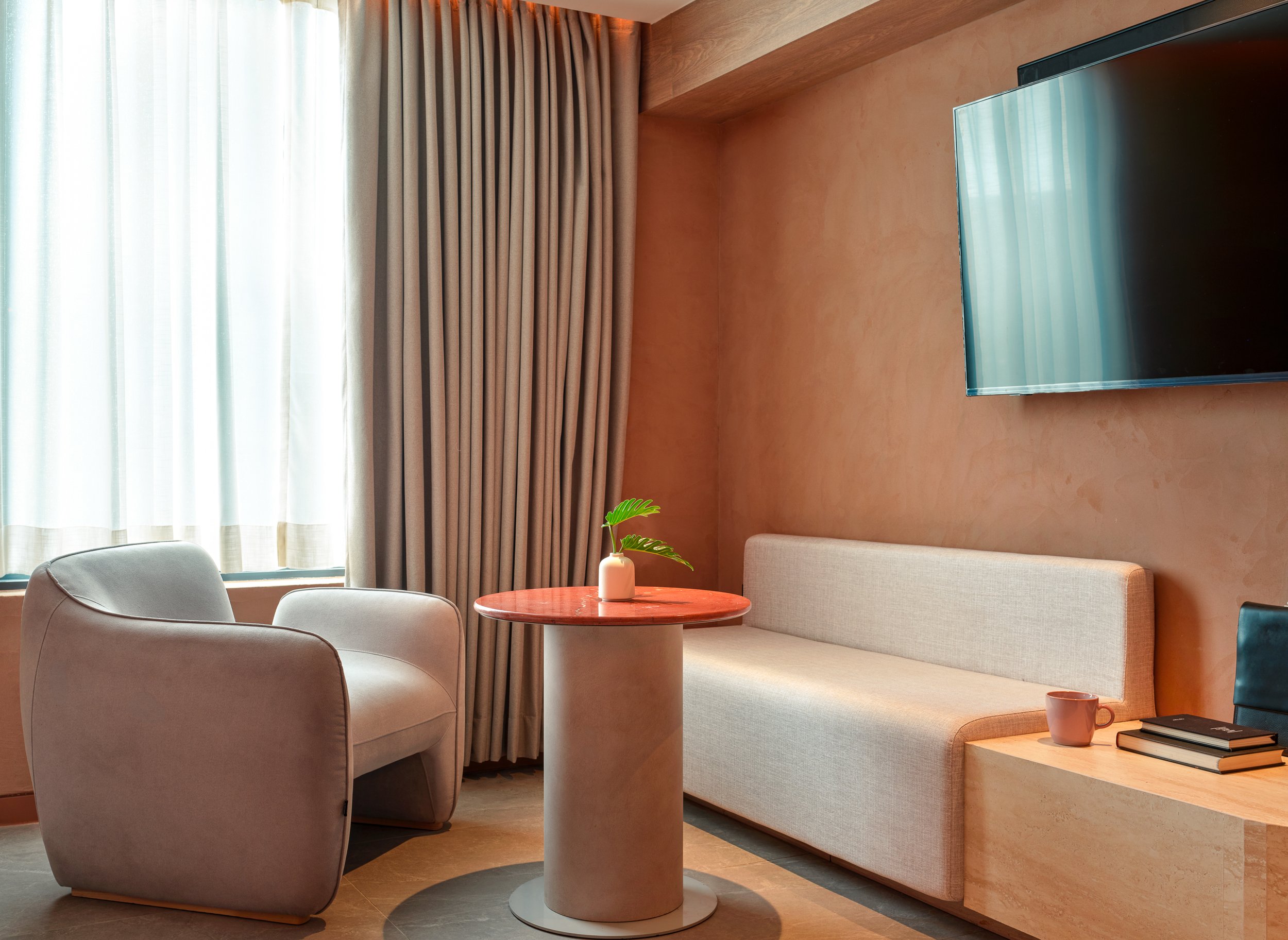 A cozy living room with neutral-colored furniture and decor. A beige armchair and a cream sofa are positioned near a small round wooden table with a white vase and green plant. Large windows with cream curtains allow natural light to fill the space, 