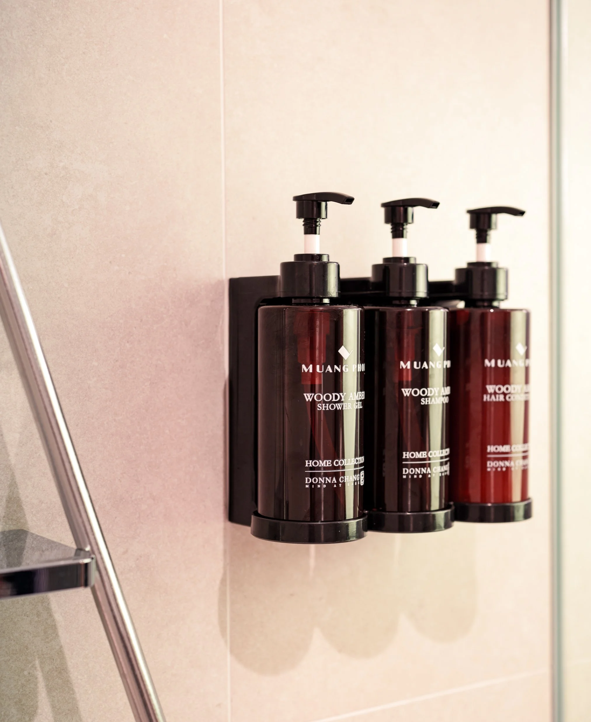 Three amber-colored bottles with black pump dispensers on a black holder on a beige wall, labeled 'Woody Amber Shower Gel', 'Woody Amber Shampoo', and 'Woody Amber Hair Cream'.