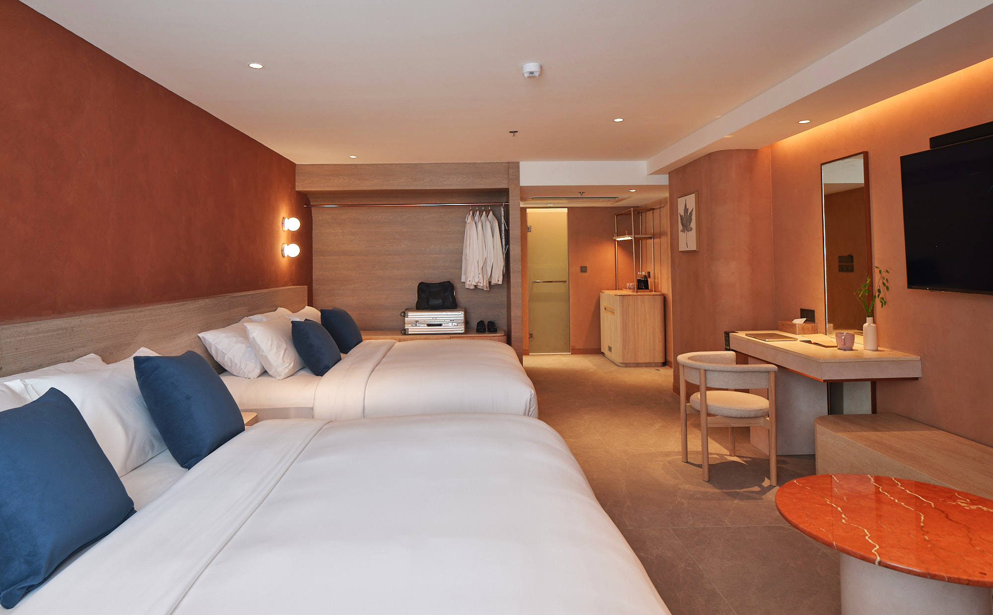 Hotel room with two beds, blue pillows, wall-mounted lights, a small closet with white robes, a black bag, a silver suitcase, a wooden desk with a mirror, chair, potted plant, wall art, flat-screen TV, marble-top table, and a door leading to the bathroom.