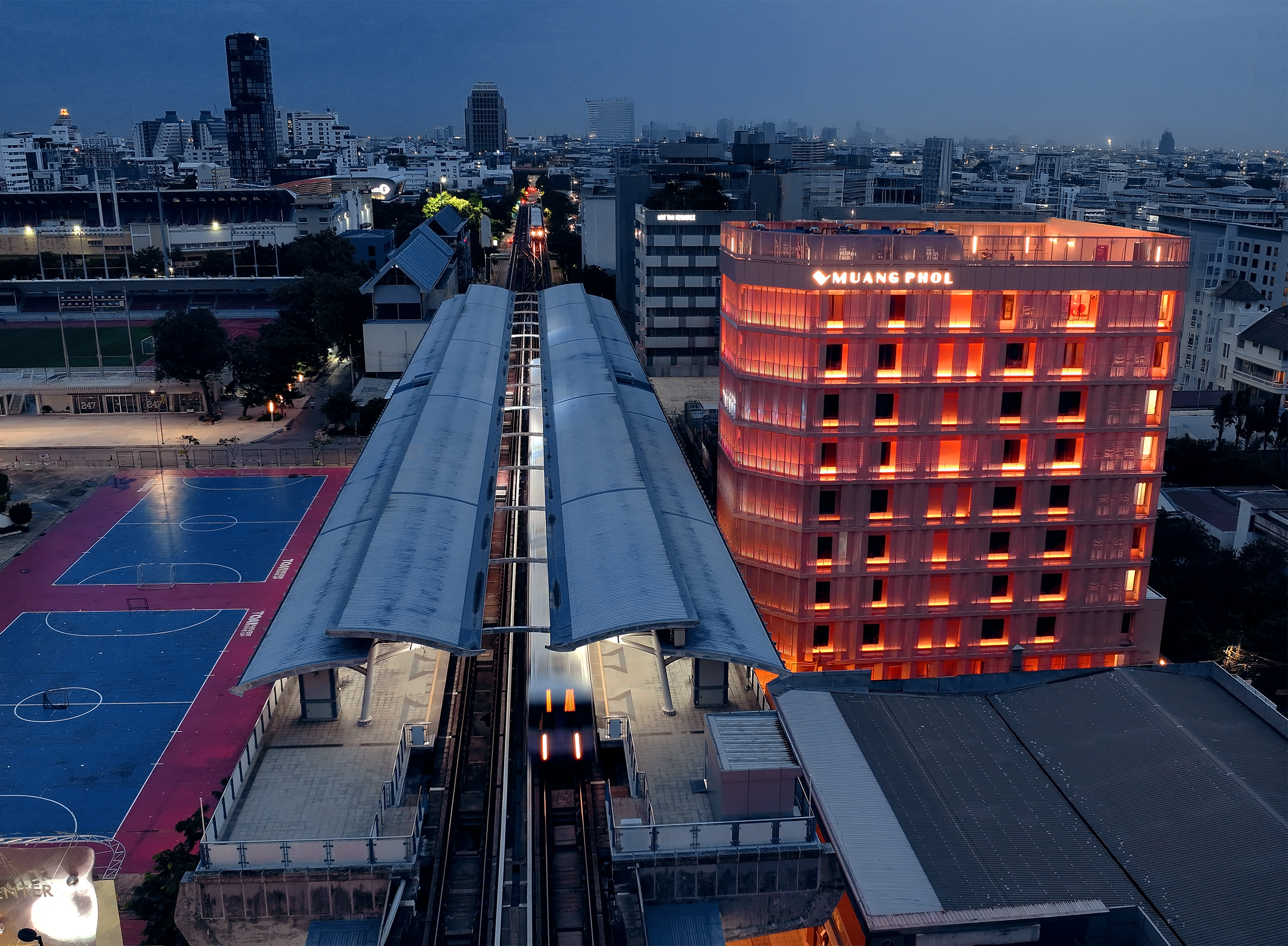 Cityscape at dusk with a modern building illuminated in orange, an elevated train station, and a sports field below.