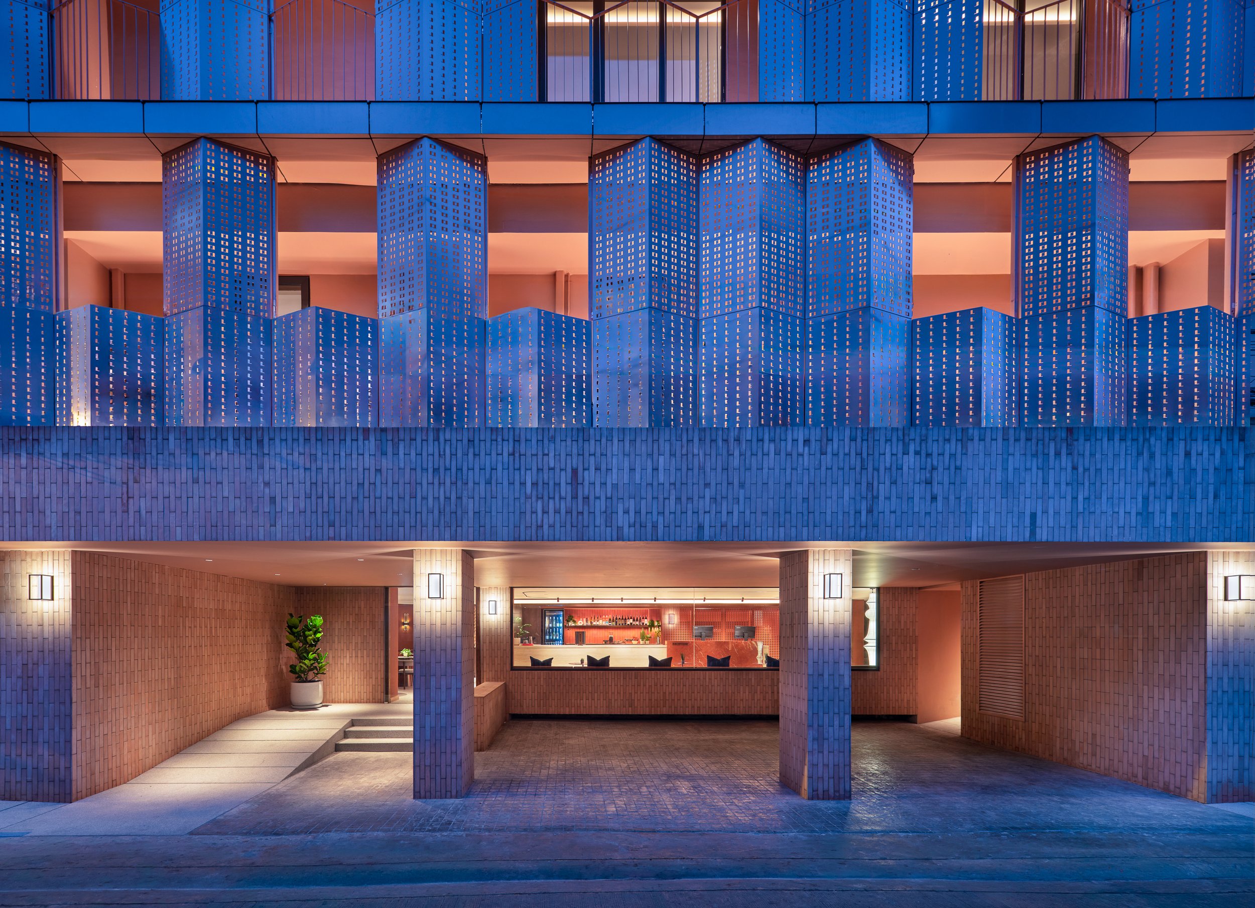 The image features a modern building facade with blue perforated panels creating geometric patterns, illuminated at dusk. The ground floor includes an open entrance area with warm interior lighting, a reception desk, and seating visible through large