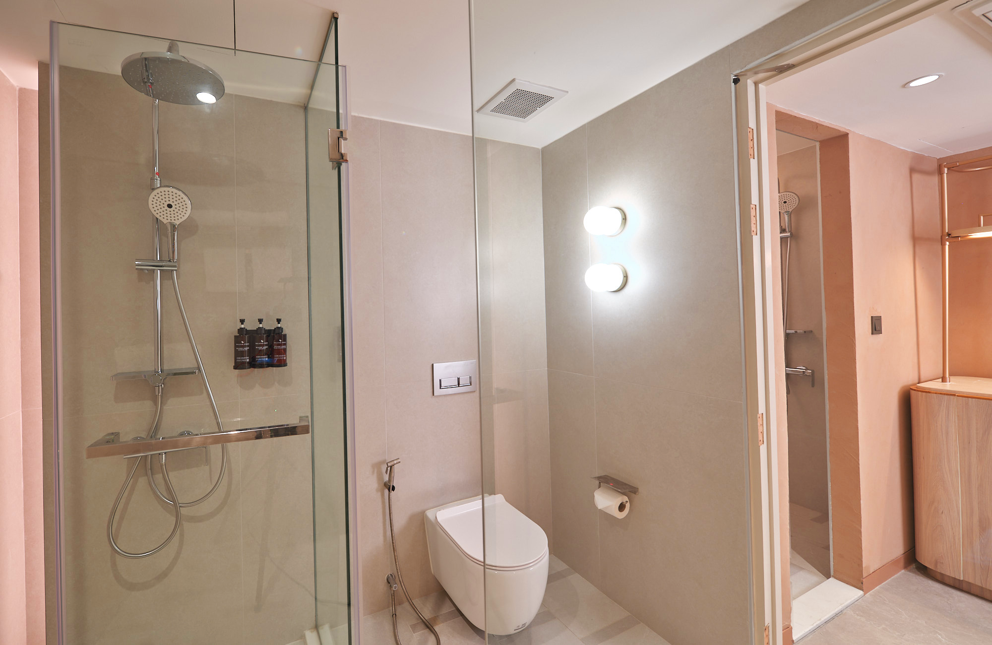 A modern bathroom with a glass shower enclosure, a ceiling-mounted rainfall showerhead, and a handheld showerhead, beige tile walls, a wall-mounted toilet, and a wall-mounted light fixture with two bulbs. There is an open doorway leading to a space with a copper pipe radiator and a wooden cabinet.