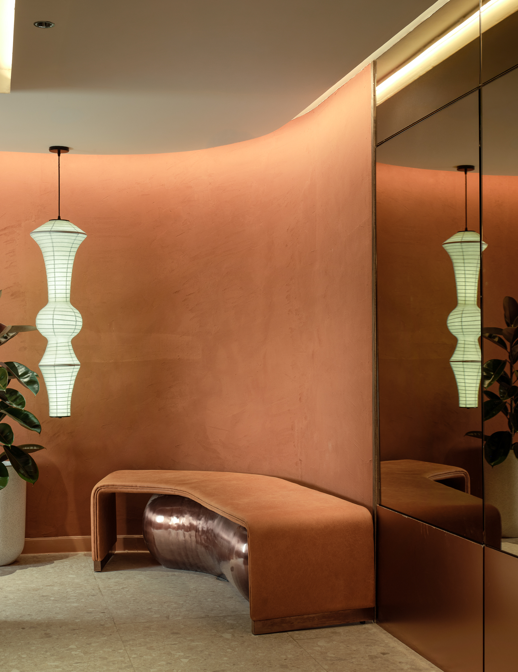 Interior space with a peach-colored cushioned seating area, a mirror on the right side, two white hanging lamps with black cords, and a green plant to the left.
