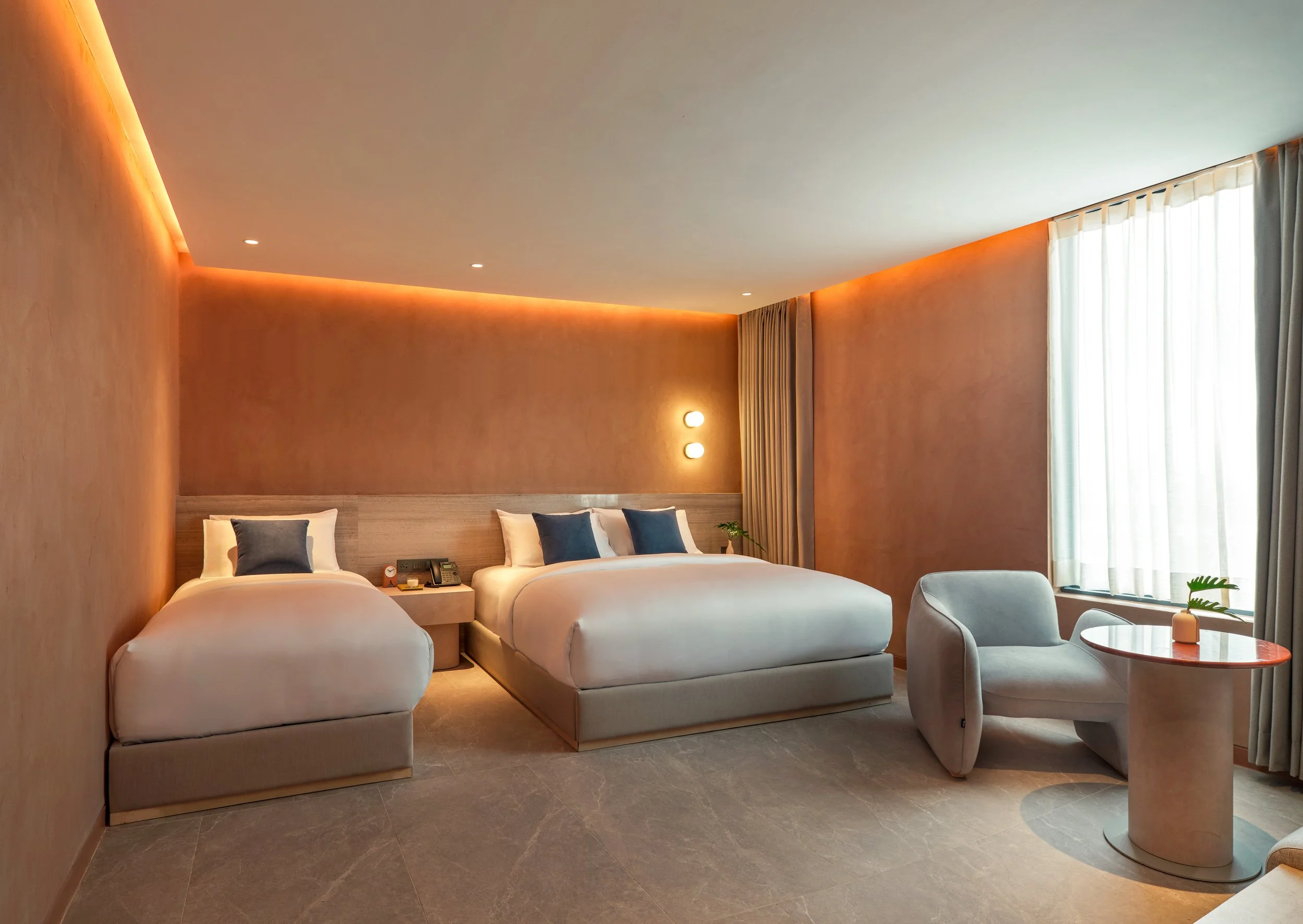 Modern hotel room with two beds, soft lighting, and a sitting area by the window.