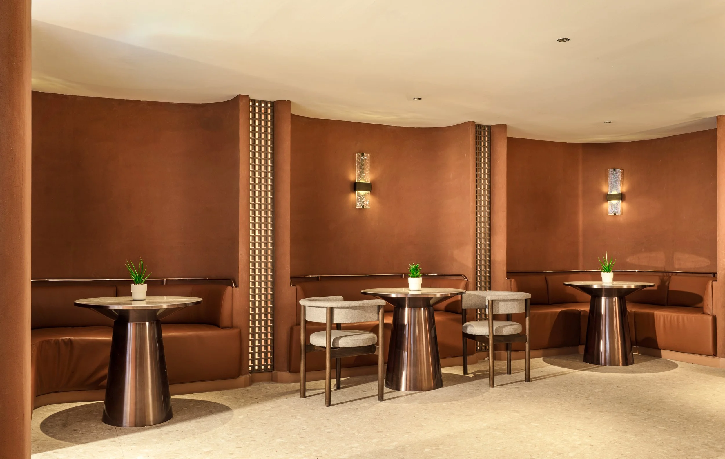 Interior of a lounge or sitting area featuring round tables with small potted plants, brown leather seating, and wall-mounted decorative lighting fixtures.