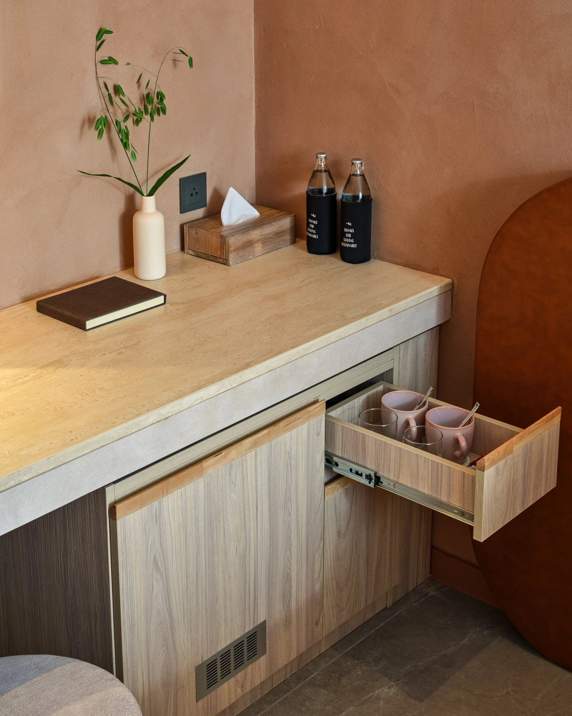 A wooden kitchen counter with a black notebook, a white tissue box, two glass bottles with black holders, pink mugs with spoons, and a potted plant on a beige wall.