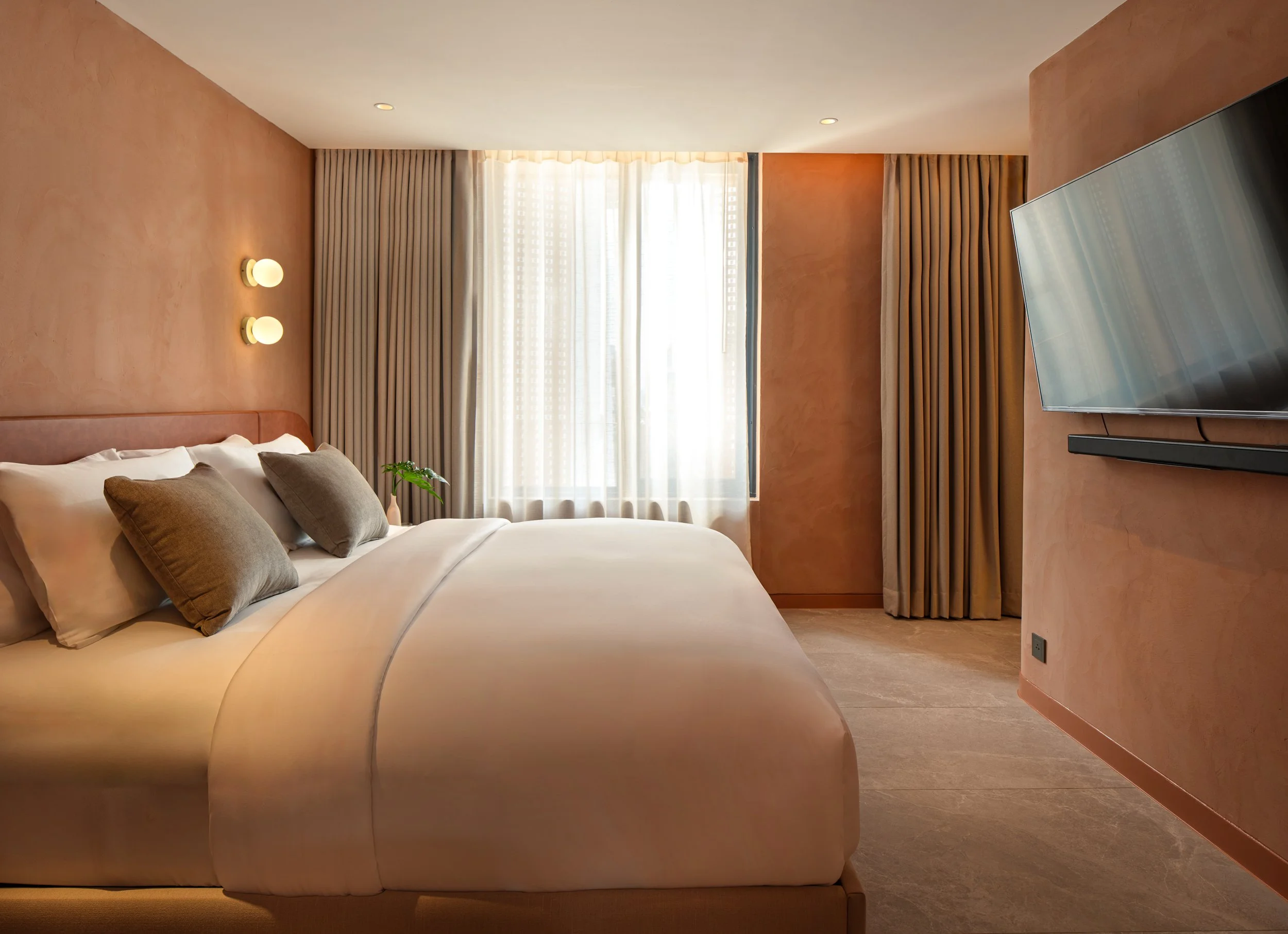 A bedroom with a bed in the foreground, beige walls, a flat-screen TV mounted on the right wall, a window with beige curtains, and wall-mounted lights above the bed.