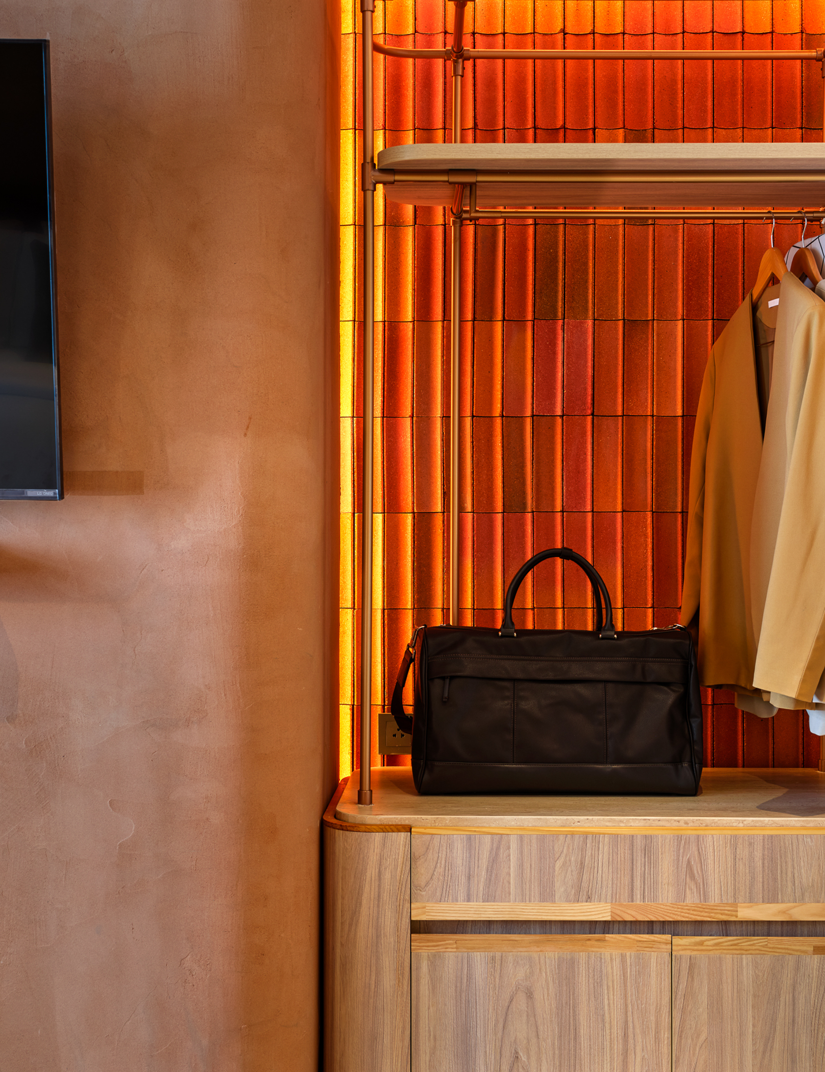 Display shelf with a black leather bag and beige clothing racked on hangers in a modern retail store, featuring a red brick wall with yellow accent lighting.
