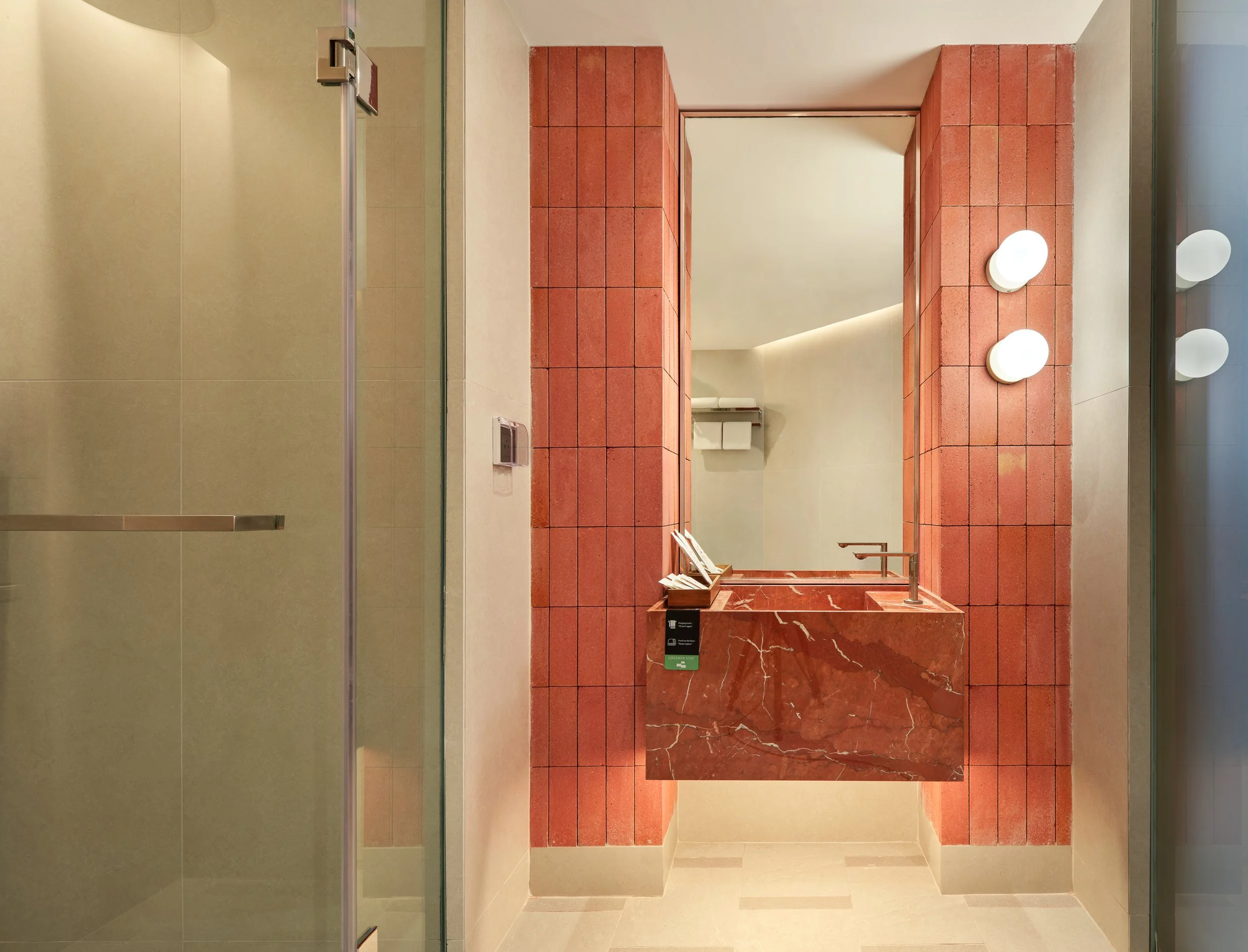 Public restroom with a red tiled wall, mirror, three white round wall lights, and a soap dispenser. The floor is beige, and there's a glass shower door on the left side.