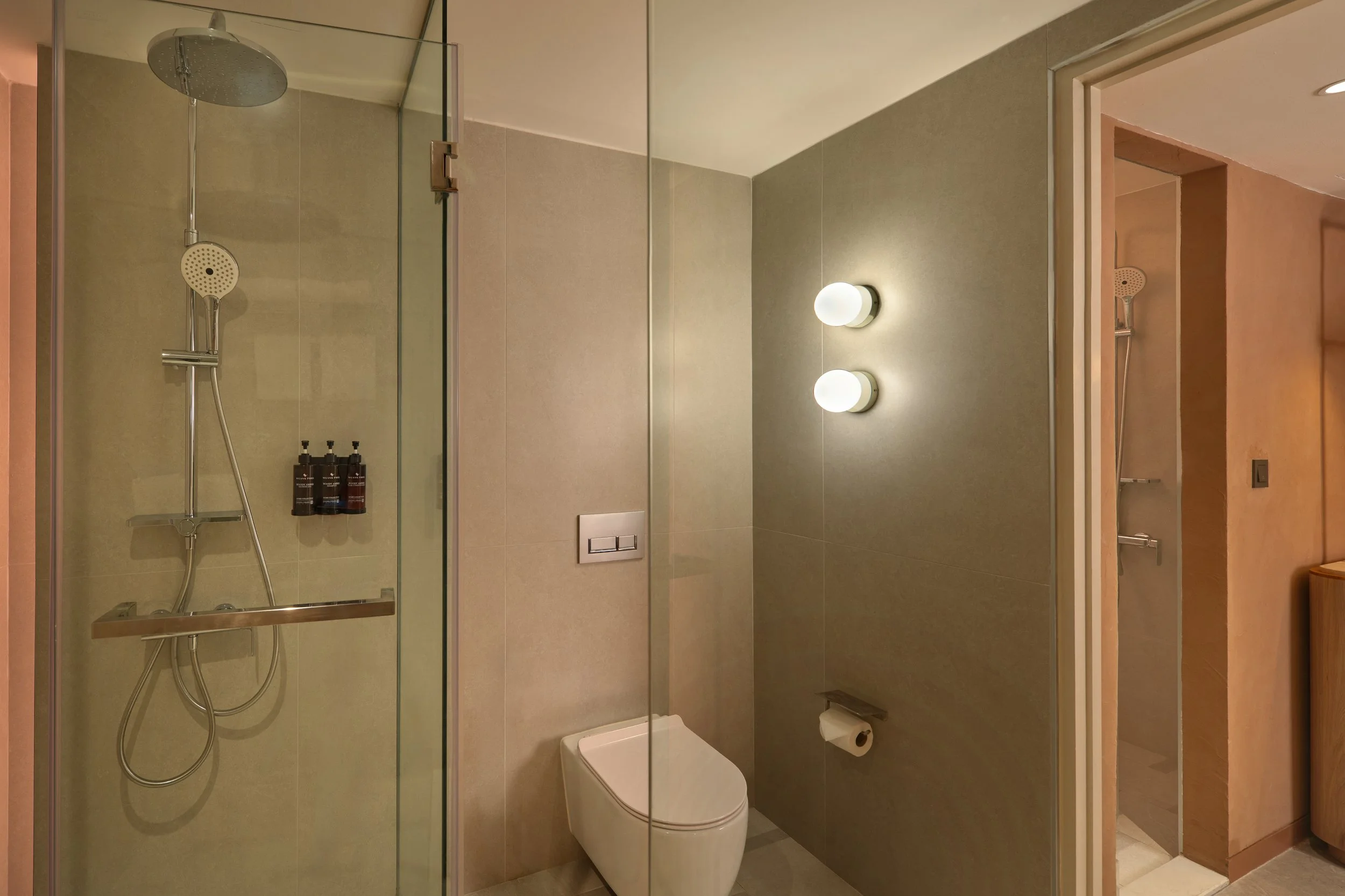 Modern bathroom with walk-in shower, wall-mounted toilet, and a large mirror. The shower has a round overhead rain shower and a handheld showerhead, with bottles of toiletries on a shelf. The lighting includes two spherical wall-mounted lights.
