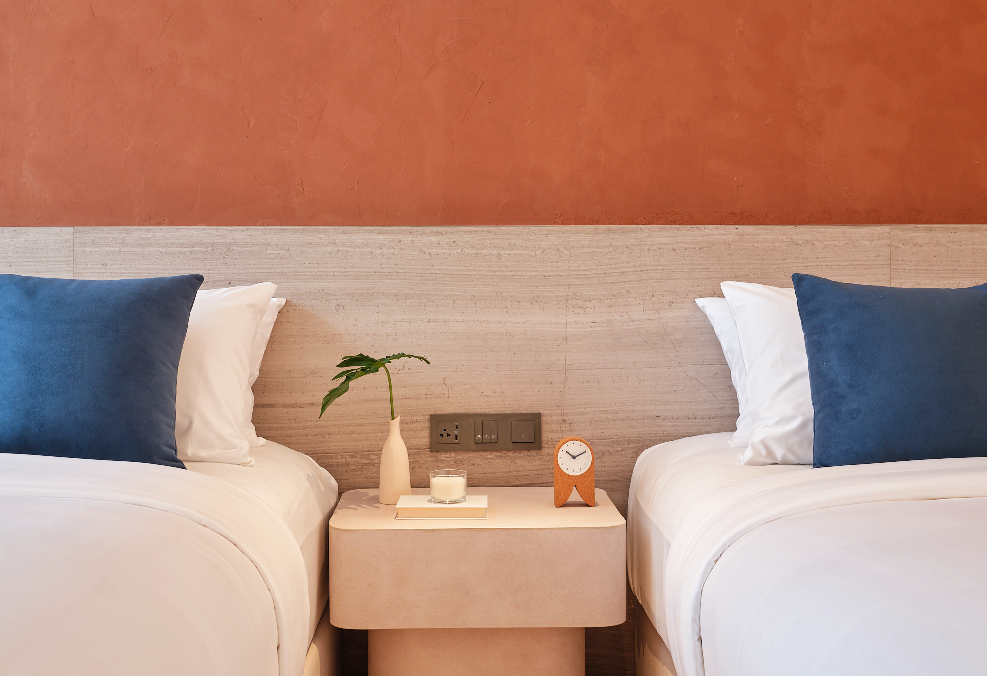 Two beds with white bedding and blue pillows, separated by a bedside table with a white vase, a glass candle holder, and a clock, against a textured terracotta wall with electrical outlets and switches.