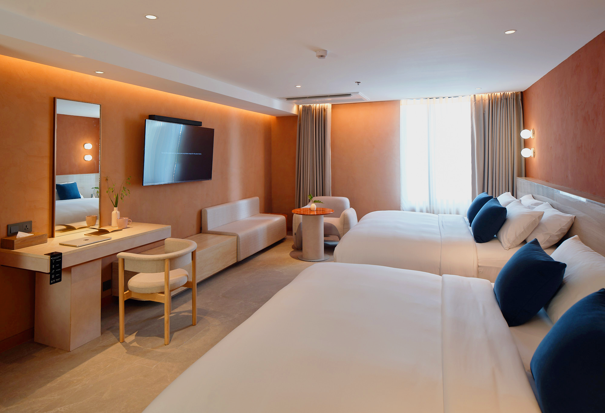 A modern hotel room with two beds, blue pillows, beige curtains, a wall-mounted TV, a mirror, a small table with a plant, and a writing desk with a chair.
