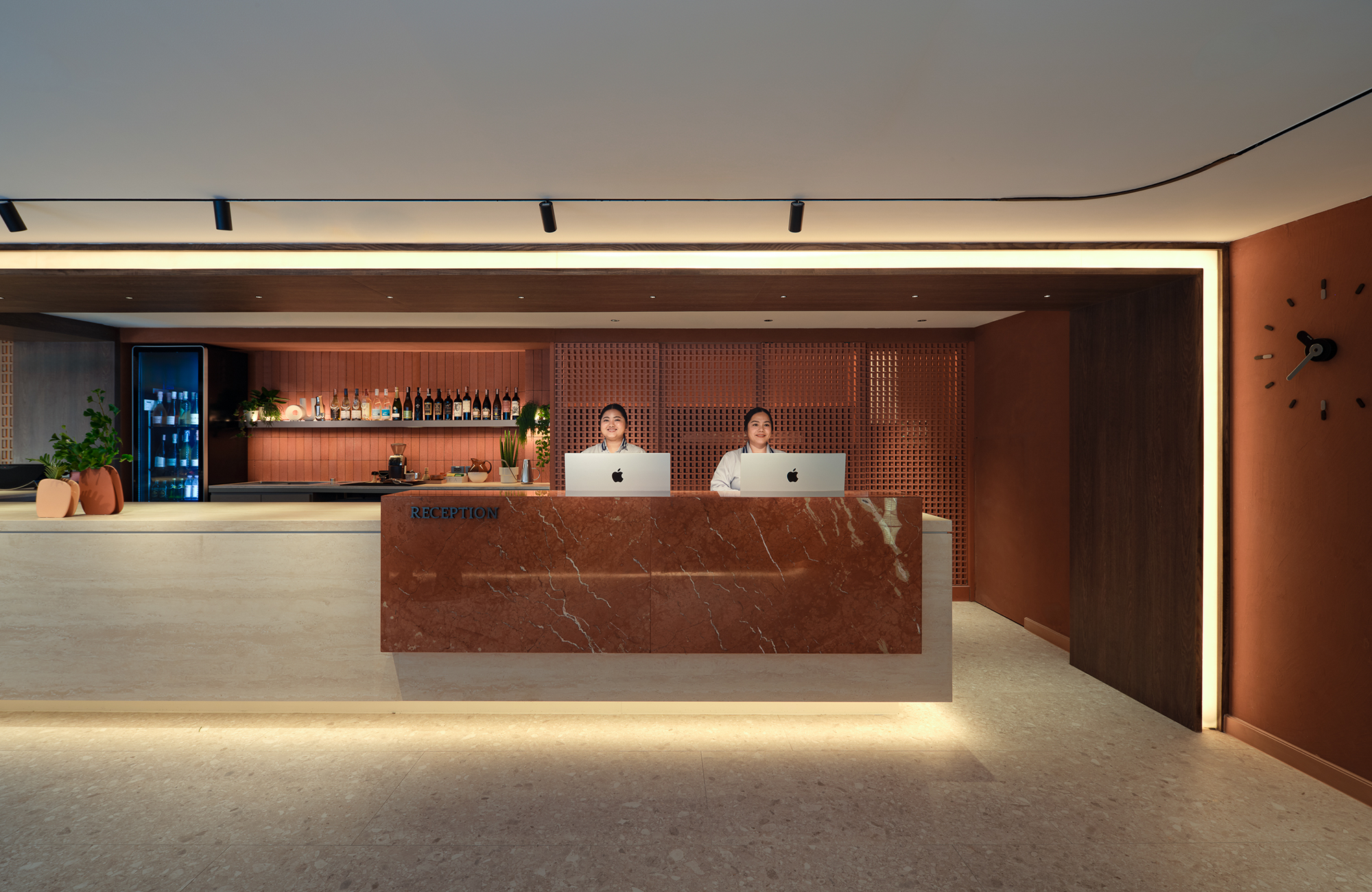 Reception area with two staff members sitting behind a marble counter, a bar with bottles and plants in the background, and modern decor with warm lighting and clock on the wall.