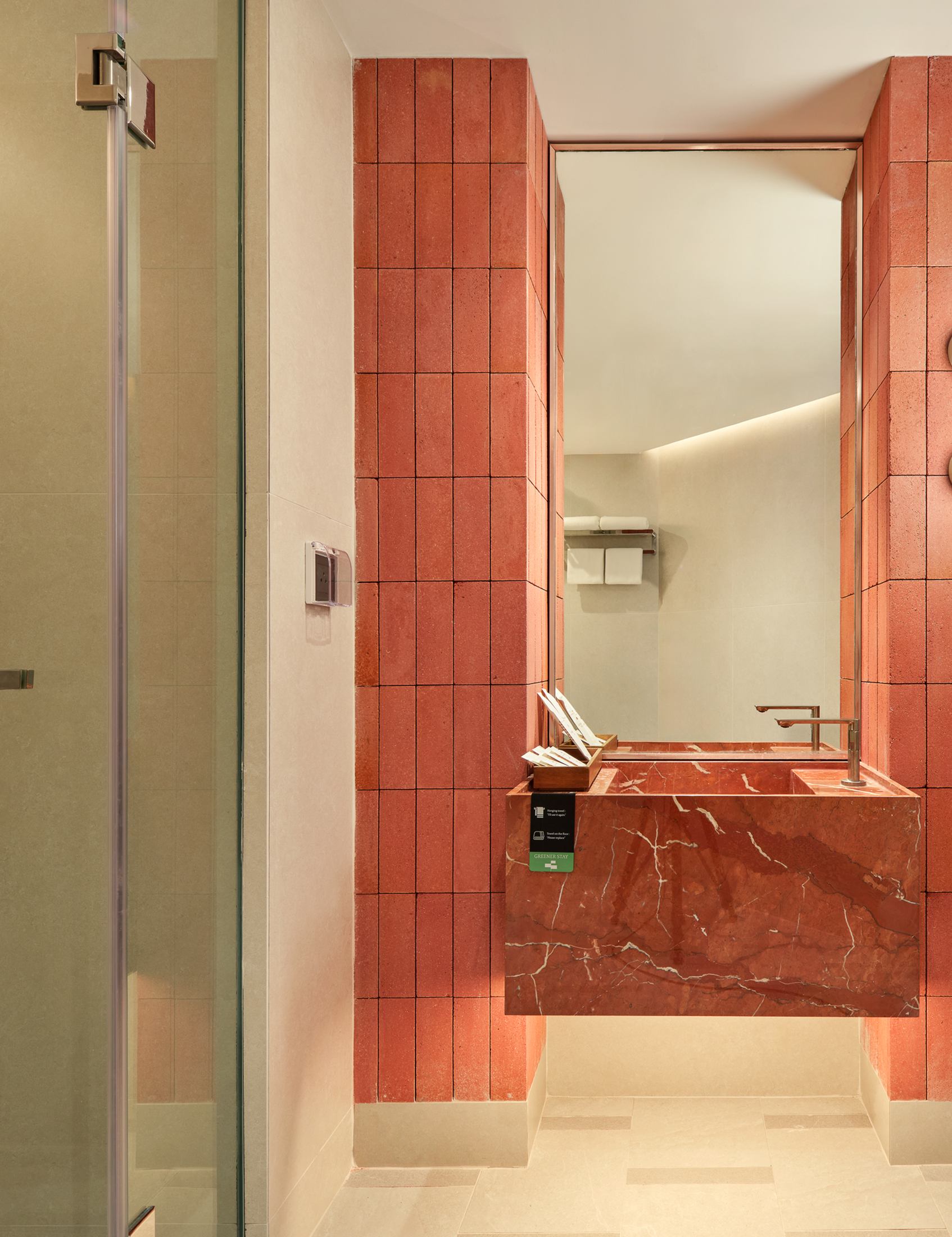 A modern bathroom with a red marble sink and backsplash, beige tiled walls, a glass shower door on the left, and a large mirror above the sink reflecting the bathroom interior.