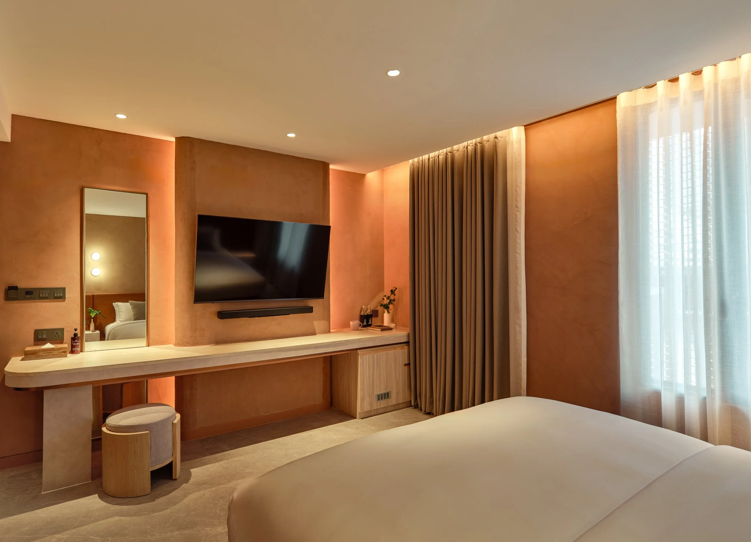 Hotel room with a wall-mounted flat-screen TV, a mirror, a window with curtains, a seating area with stools, and part of a bed.