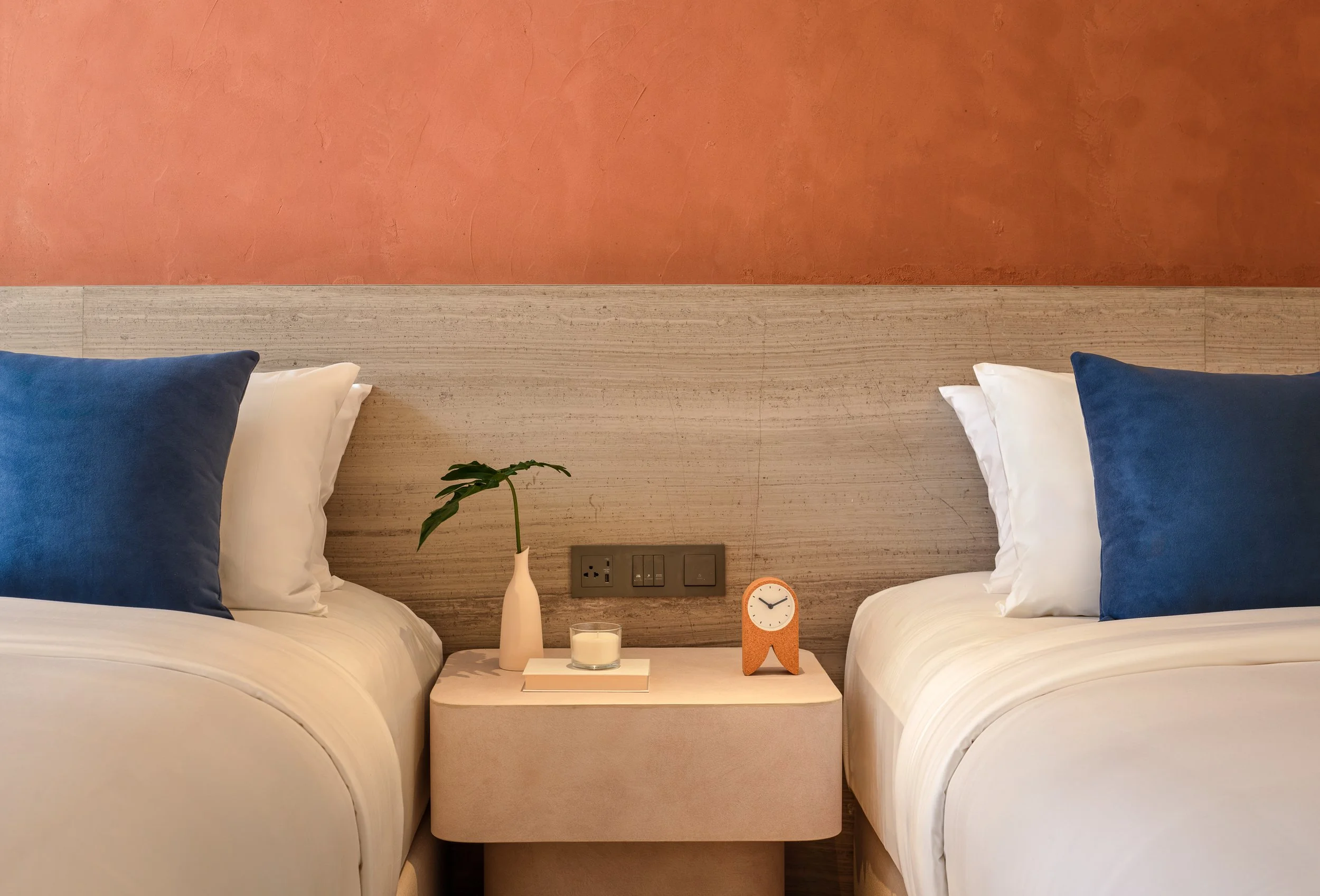 A bedroom with two beds, each with white and navy pillows, a beige headboard, a small nightstand with a vase, a clock, and candles, against a burnt orange textured wall with visible cracks.
