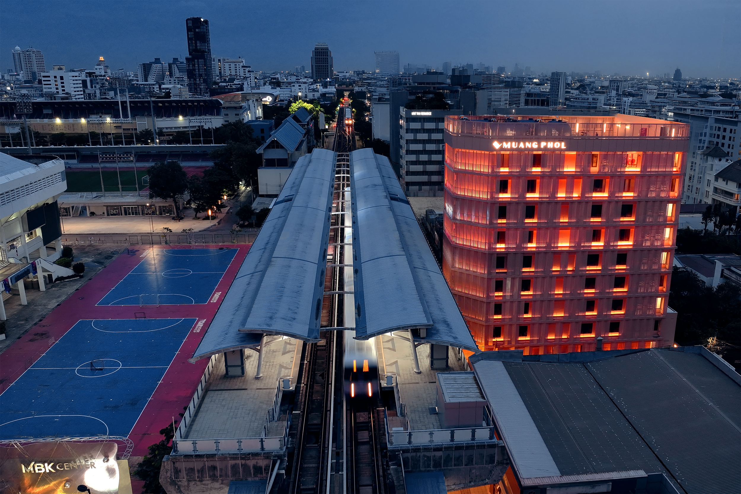 A cityscape at dusk with a train on an elevated track, a building with orange lighting labeled 'MUANG PHOL,' sports courts, and a stadium in the background.