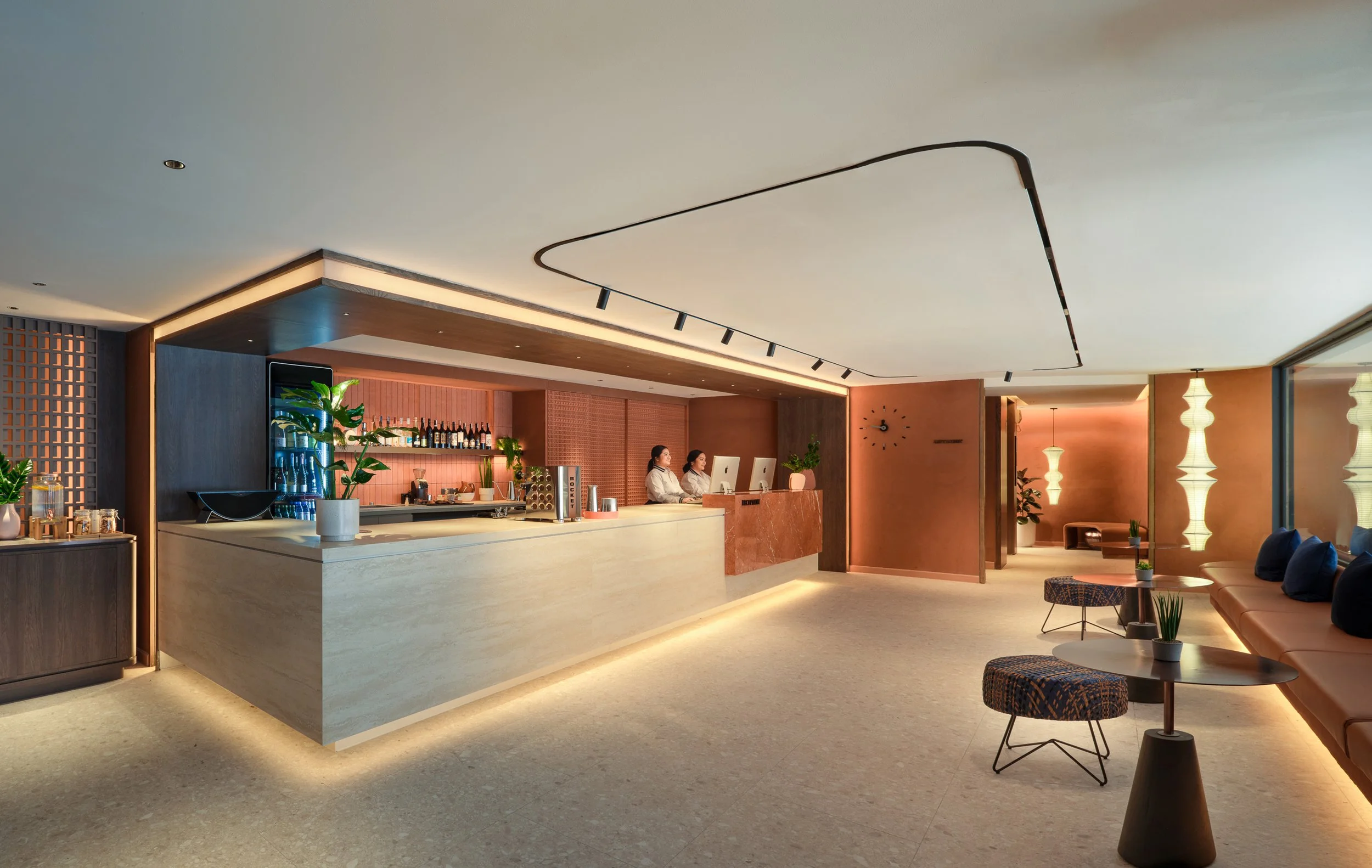 Modern hotel lobby with a reception desk, two staff members working on computers, indoor plants, and seating area with chairs and a sofa, illuminated with warm lighting.